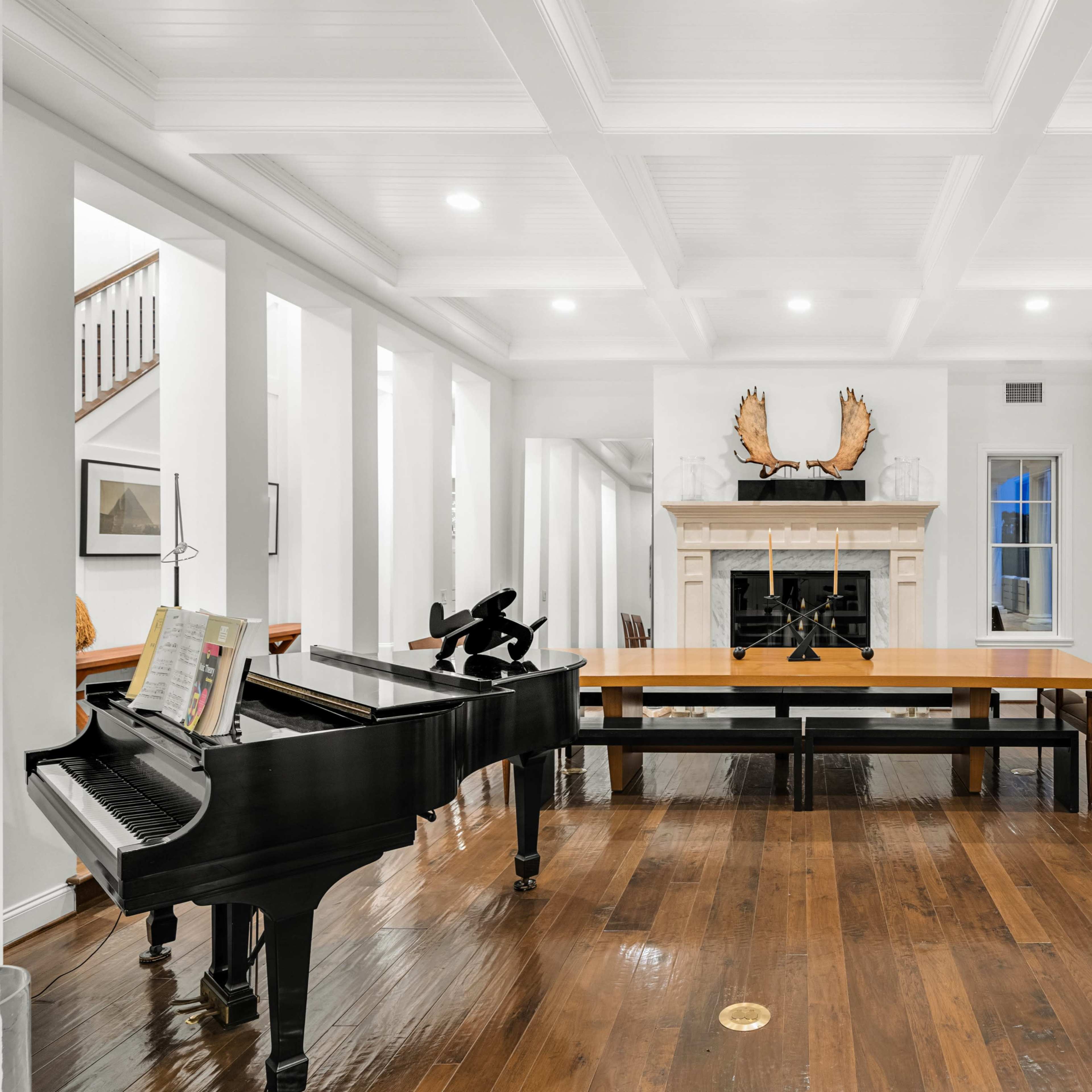 A spacious room features a grand piano, a large dining table, and a stone fireplace with antler decor.