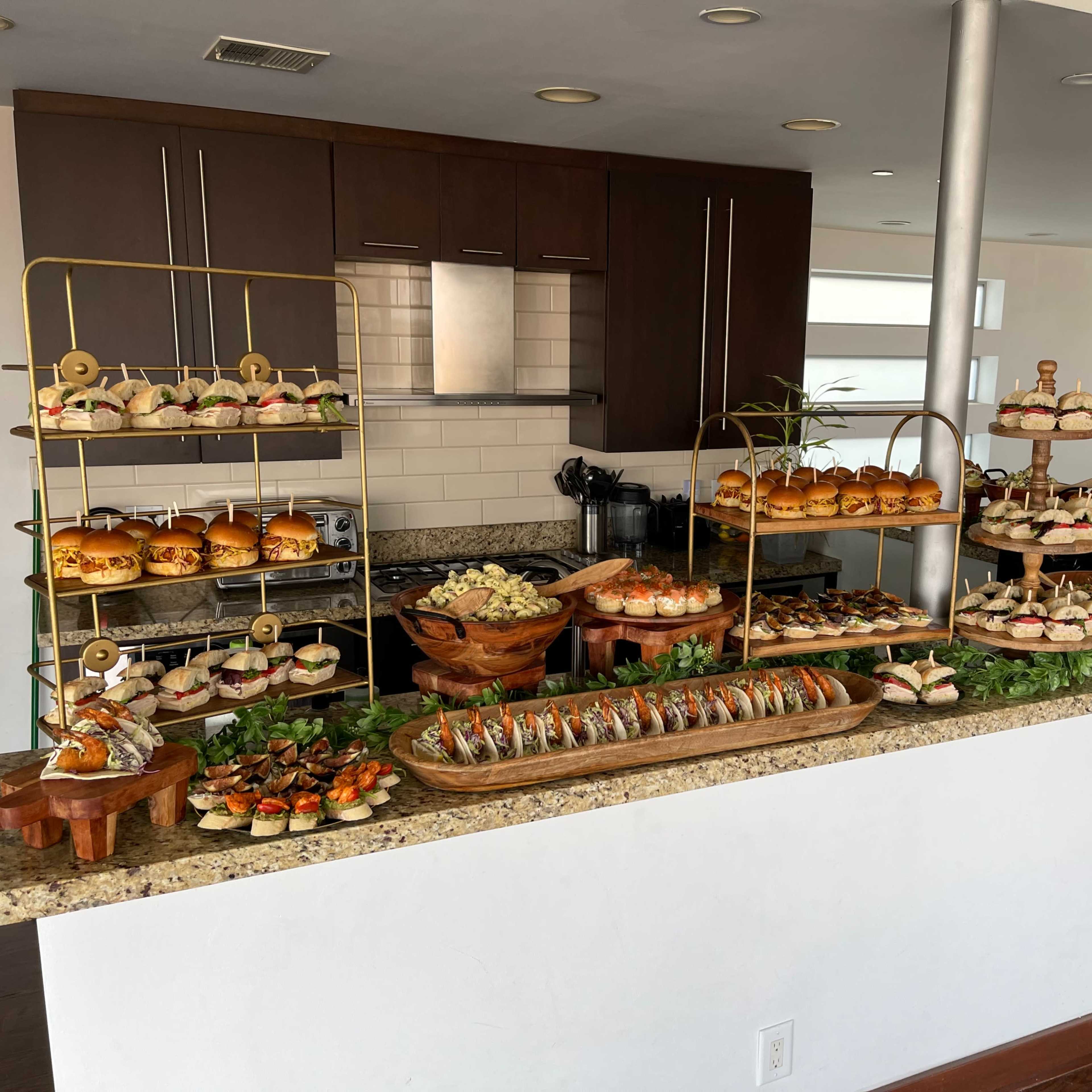 A buffet table is set up with various platters of sandwiches, sushi, and desserts, organized on metal and wooden stands in a modern kitchen.