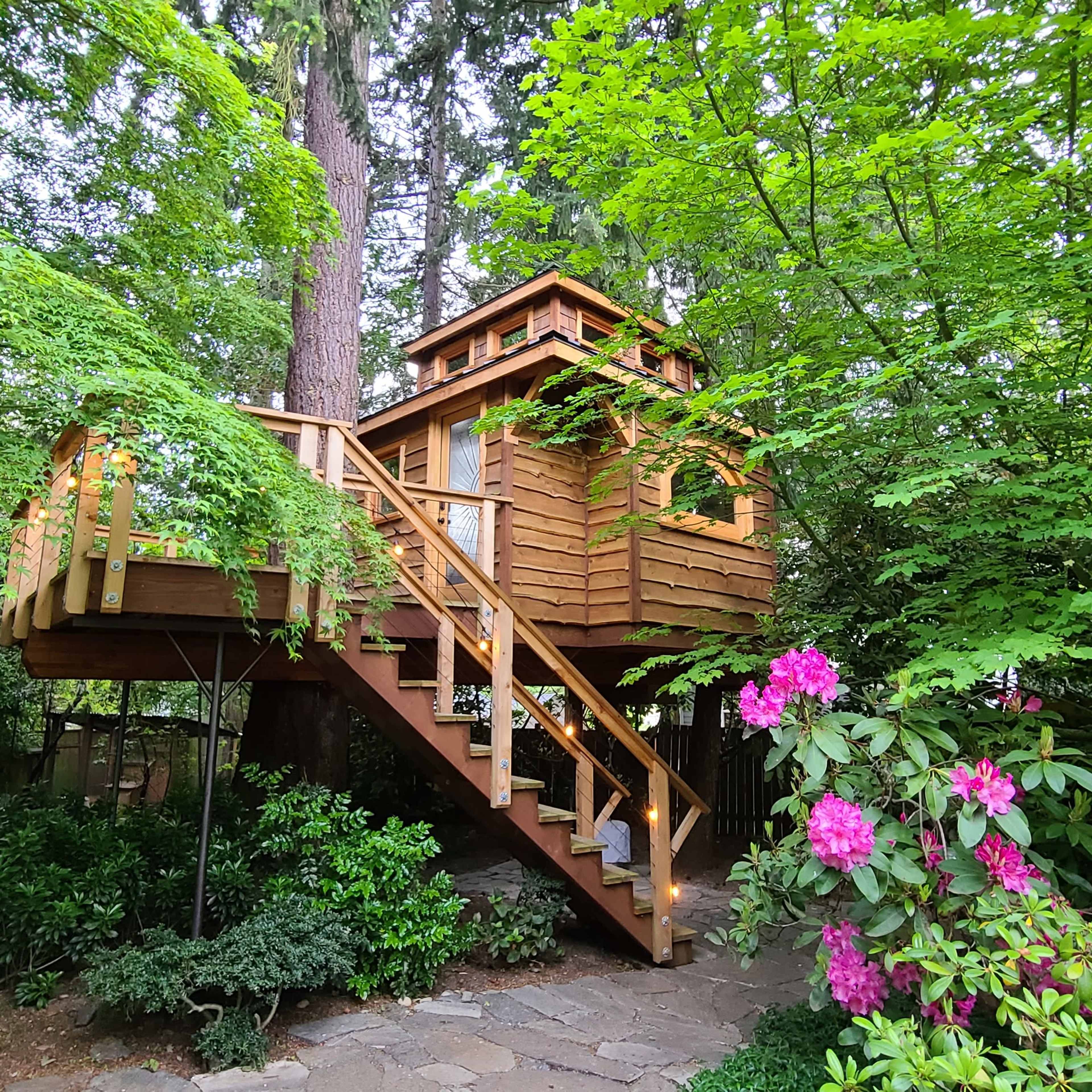 A wooden treehouse is elevated on a platform surrounded by lush greenery and colorful flowers.