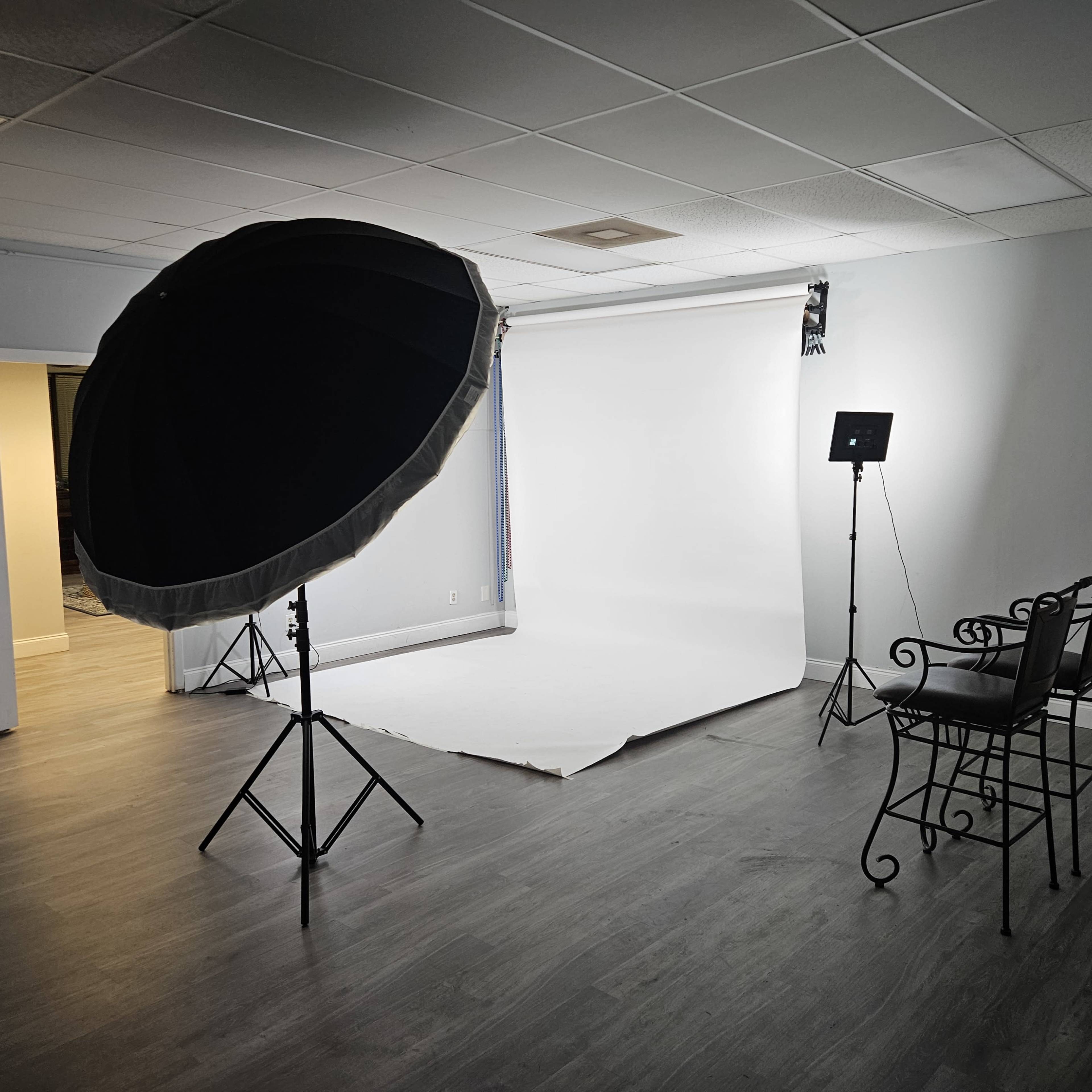 A photography studio featuring a softbox light, a spotlight, a backdrop, and a pair of metal chairs arranged on a wooden floor.