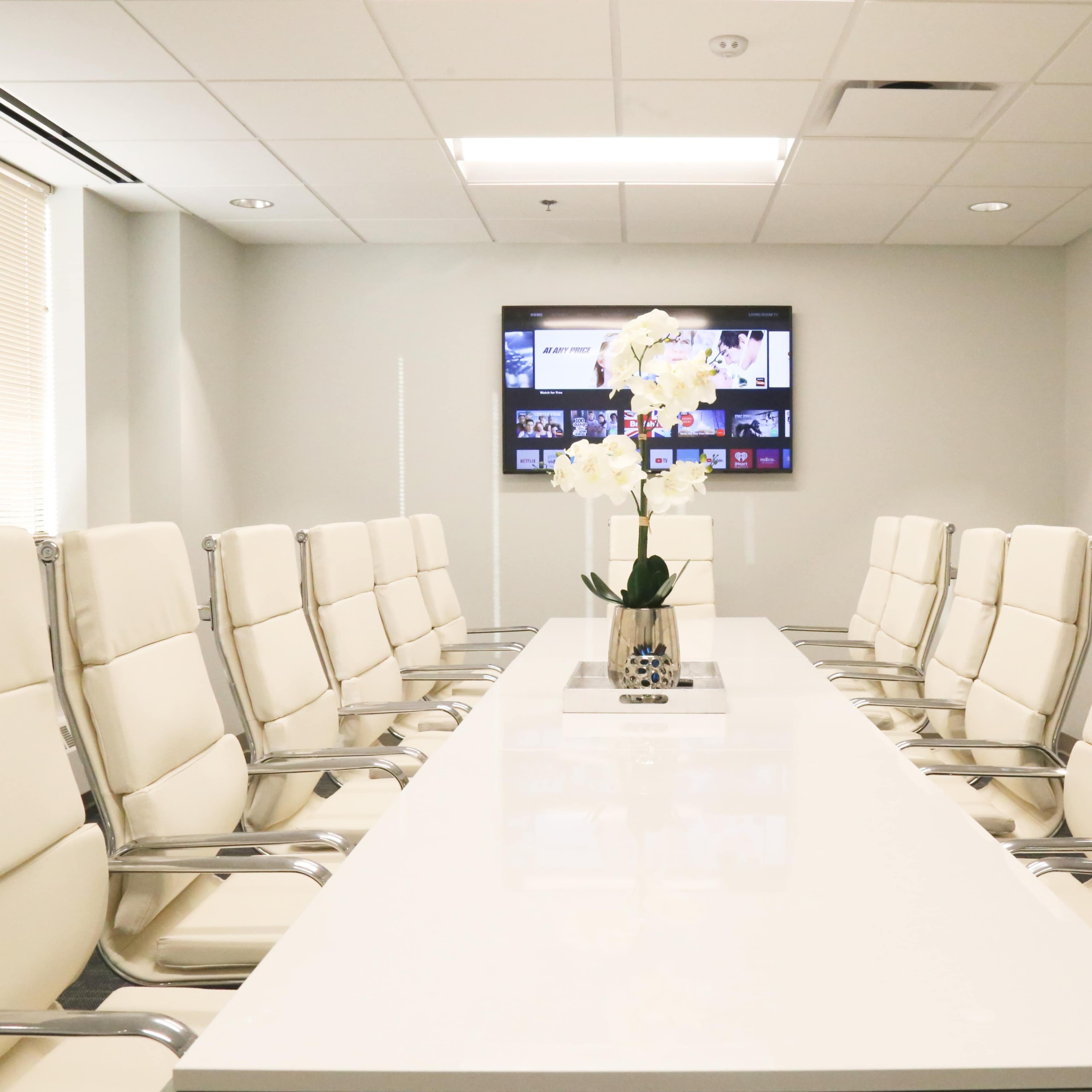 A modern conference room features a long white table surrounded by ten white leather chairs, with a television display on the wall.
