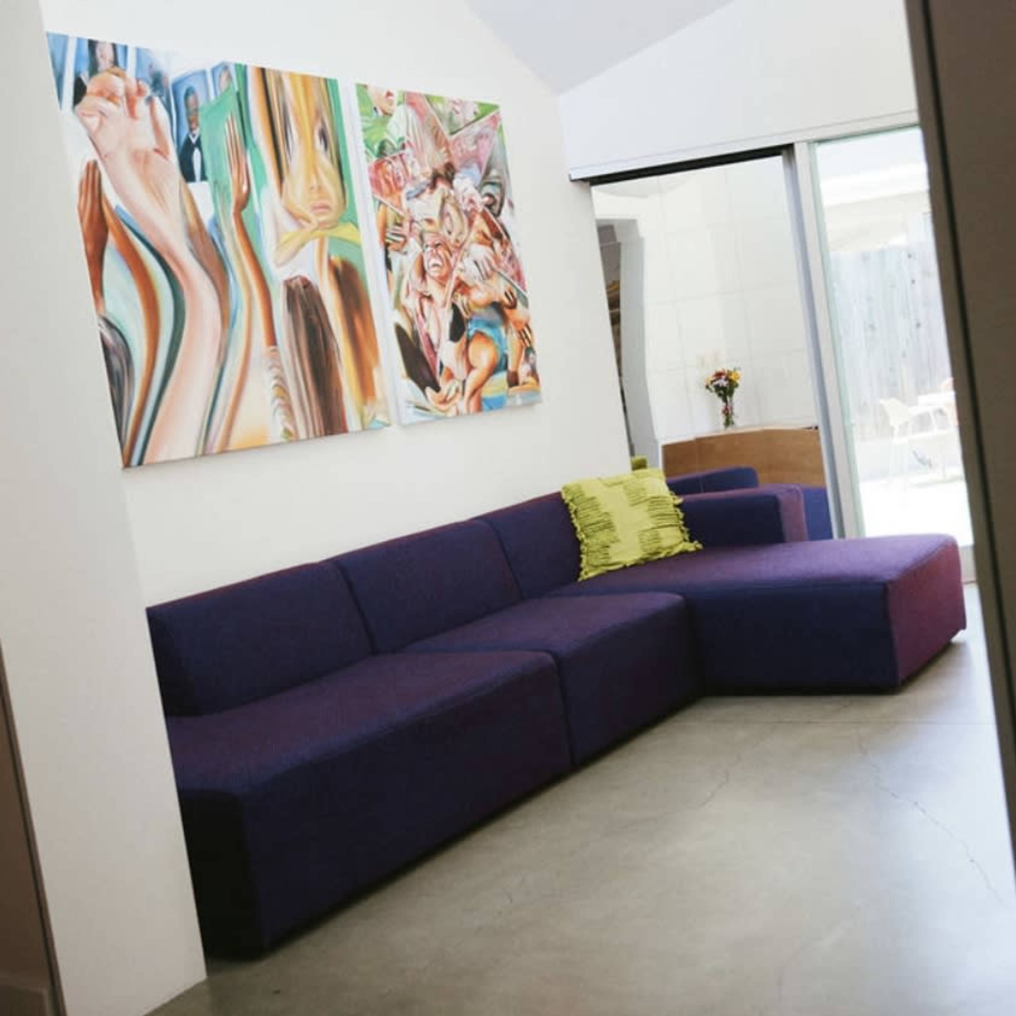 A modern living room with a purple sectional sofa against a white wall adorned with colorful abstract paintings.