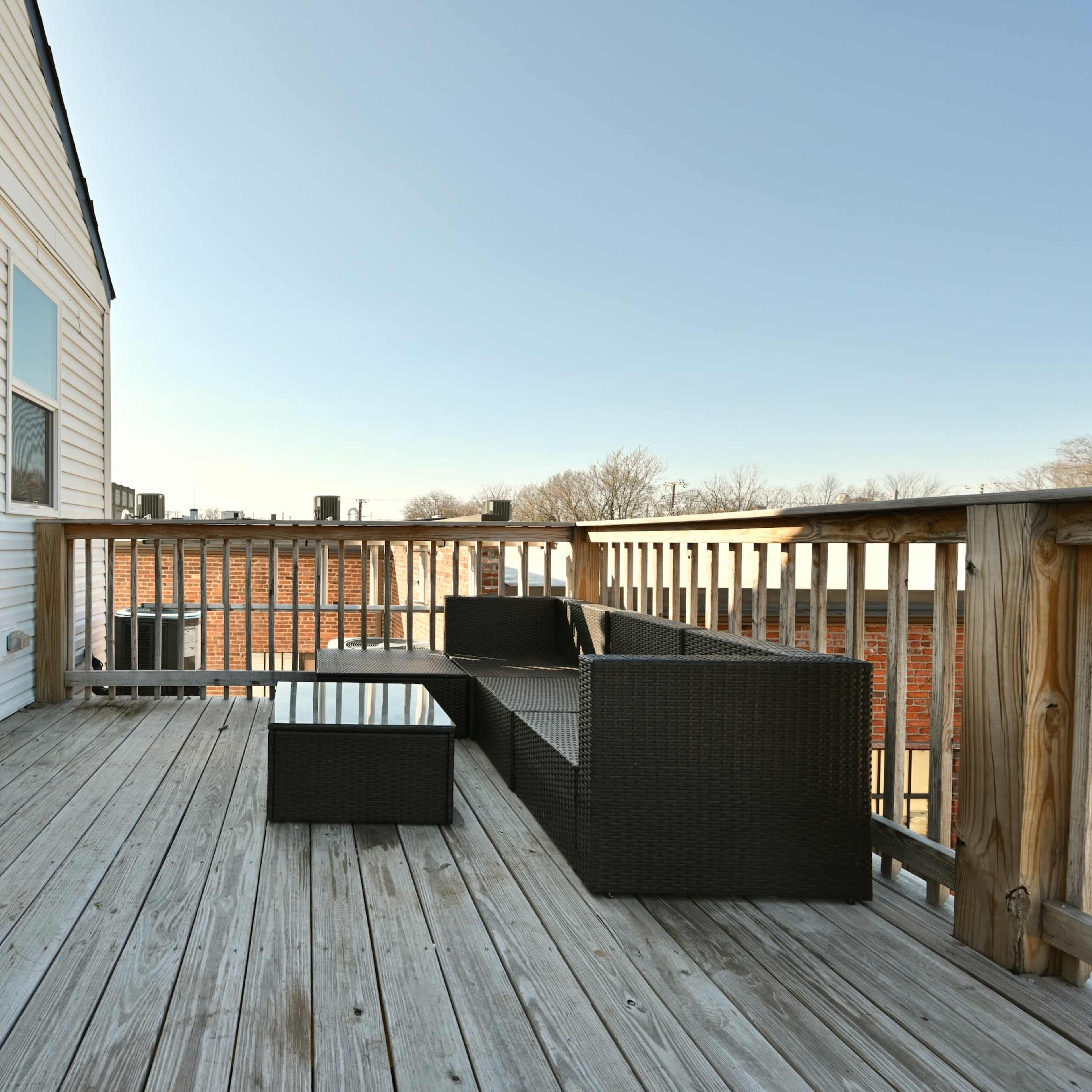 A wooden deck features a seating area with a dark wicker sofa and a glass-top coffee table, adjacent to a sliding glass door.