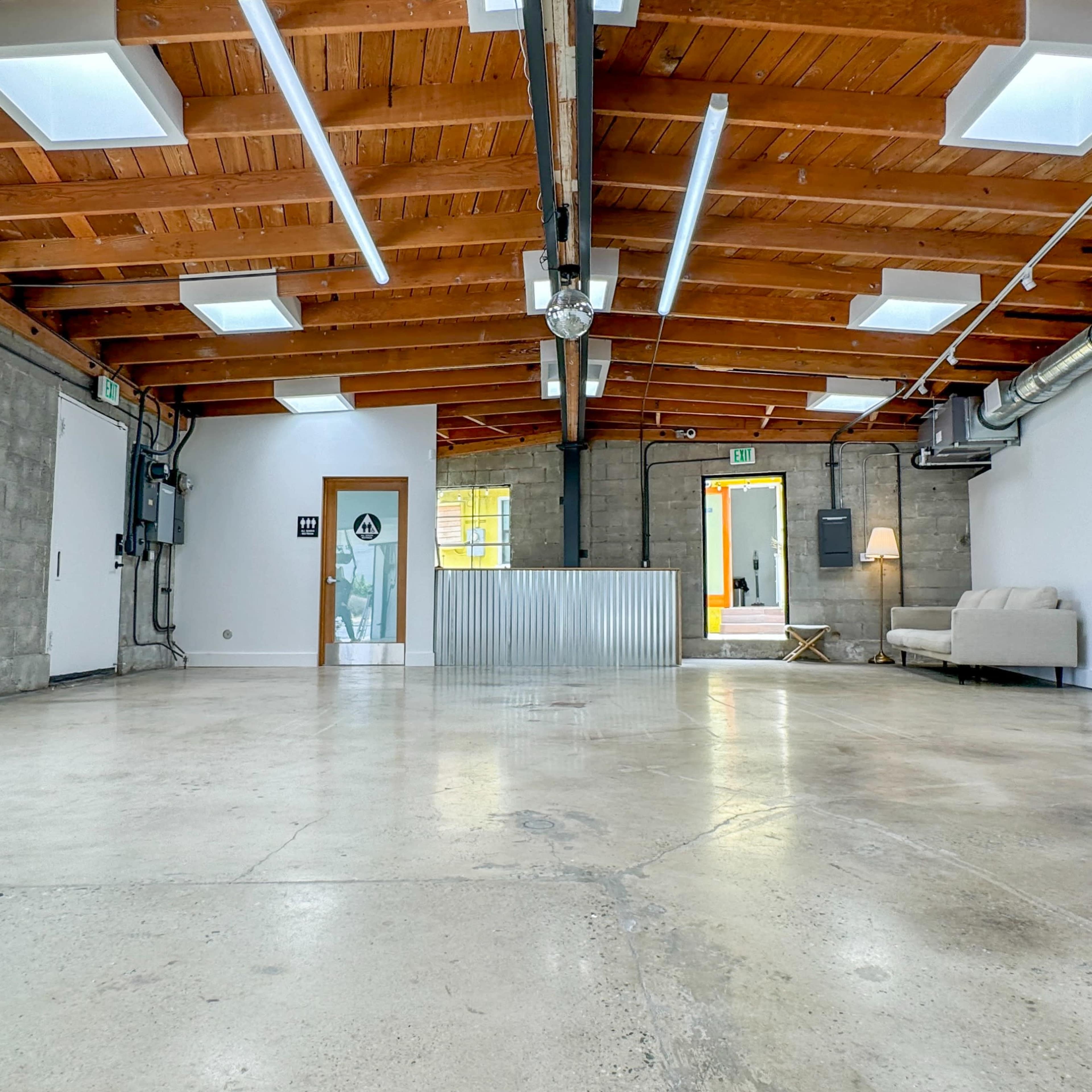 A spacious, empty industrial-style room with concrete floors, wooden beams, large windows, and minimal furnishings.