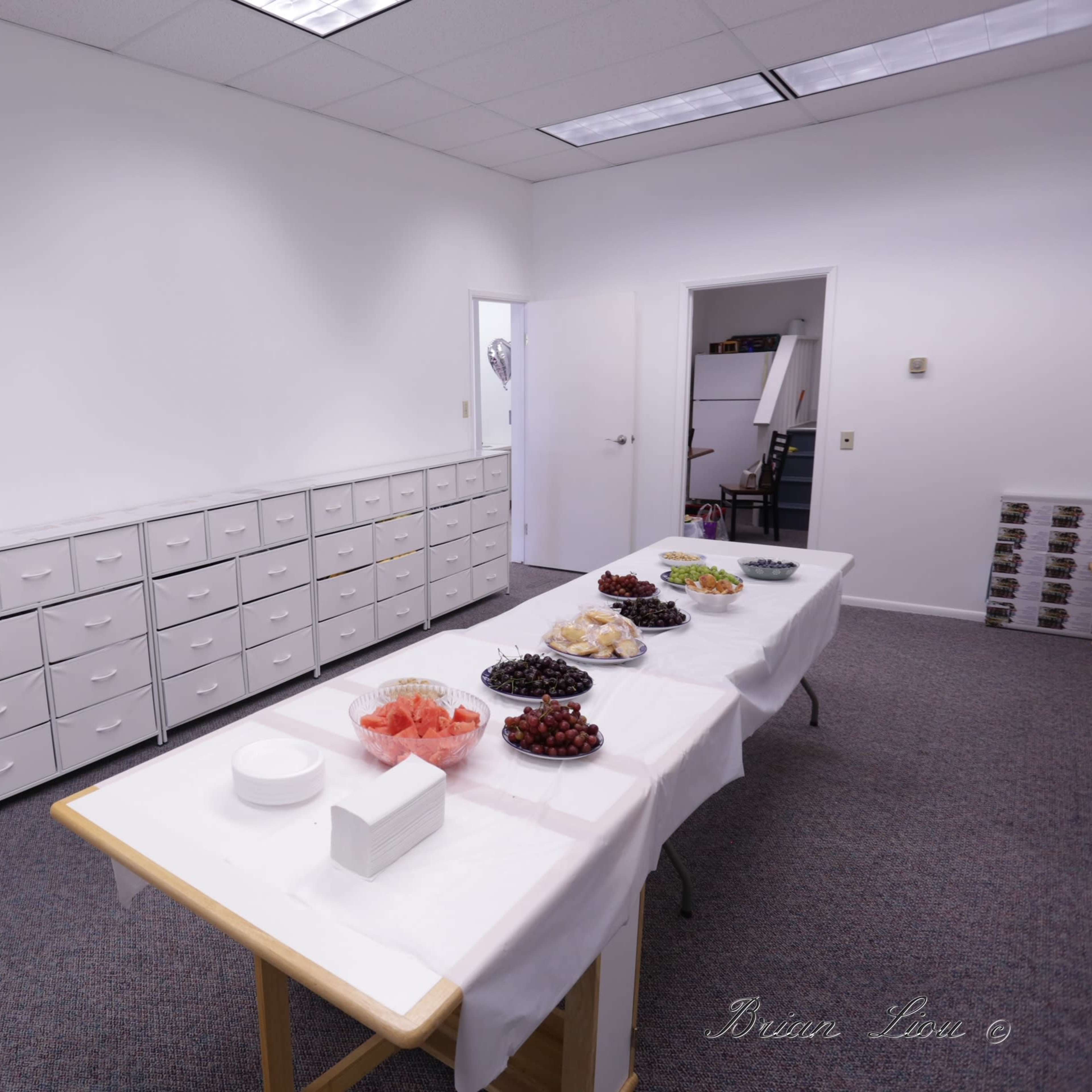 A spacious room with white walls, featuring long tables set with various bowls of fruit and a row of cabinets lining one side.