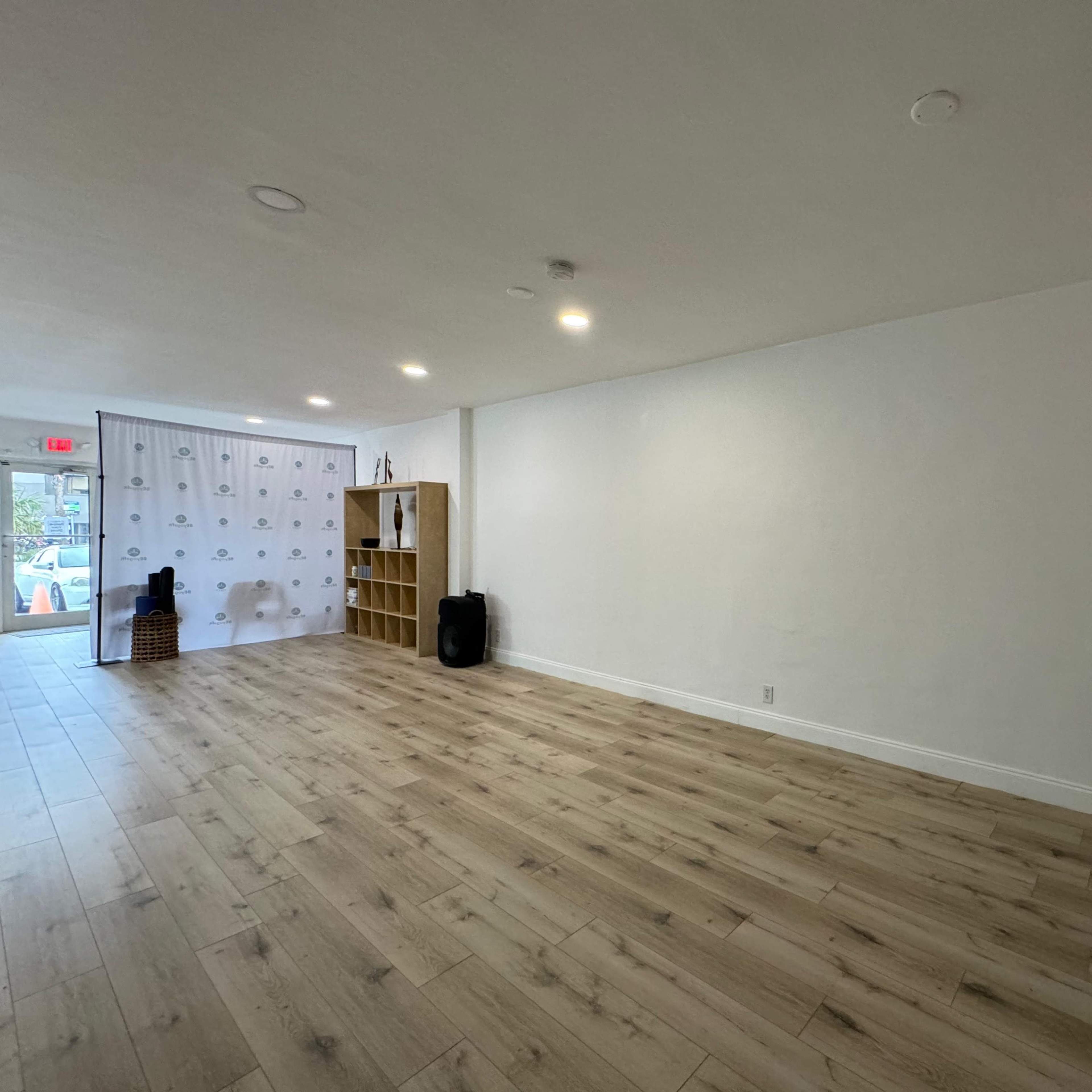 A spacious, empty room with light-colored wooden flooring and a white wall featuring a refrigerator and a shelf against one side.
