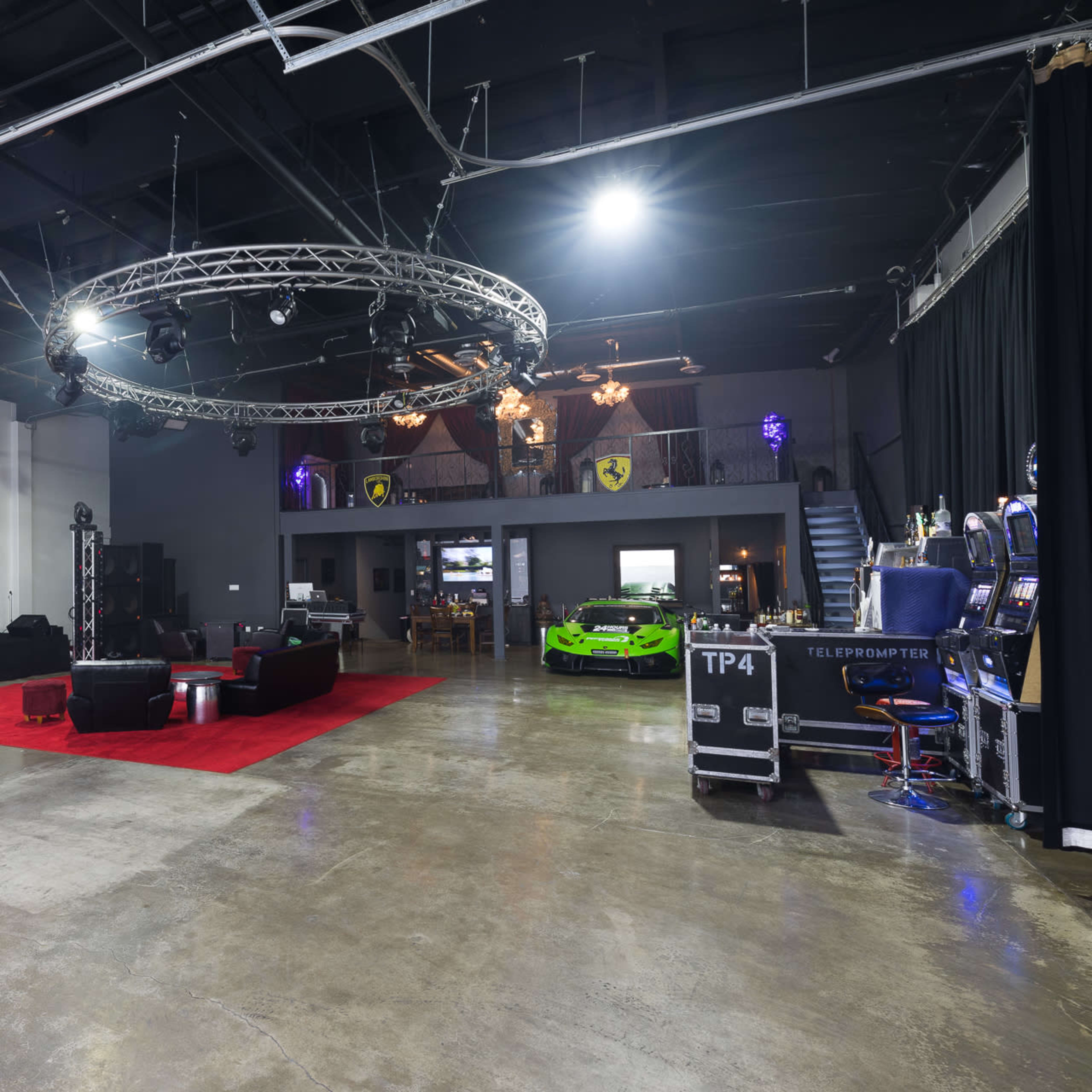 The spacious interior features a polished concrete floor, stage lighting, seating area with black couches, a green sports car, and entertainment equipment.