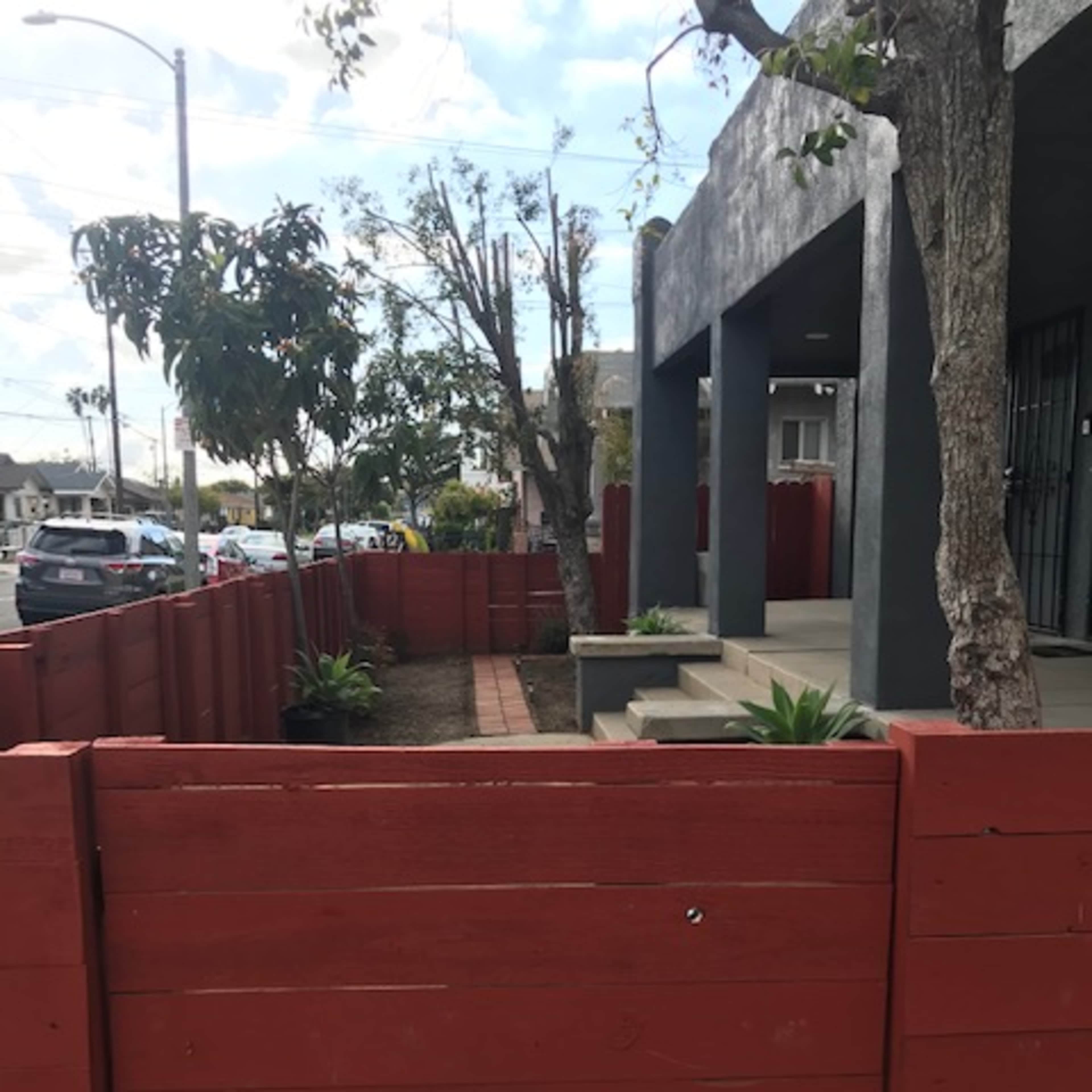 A red fenced area with potted plants and a walkway next to a building, set against a partly cloudy sky.