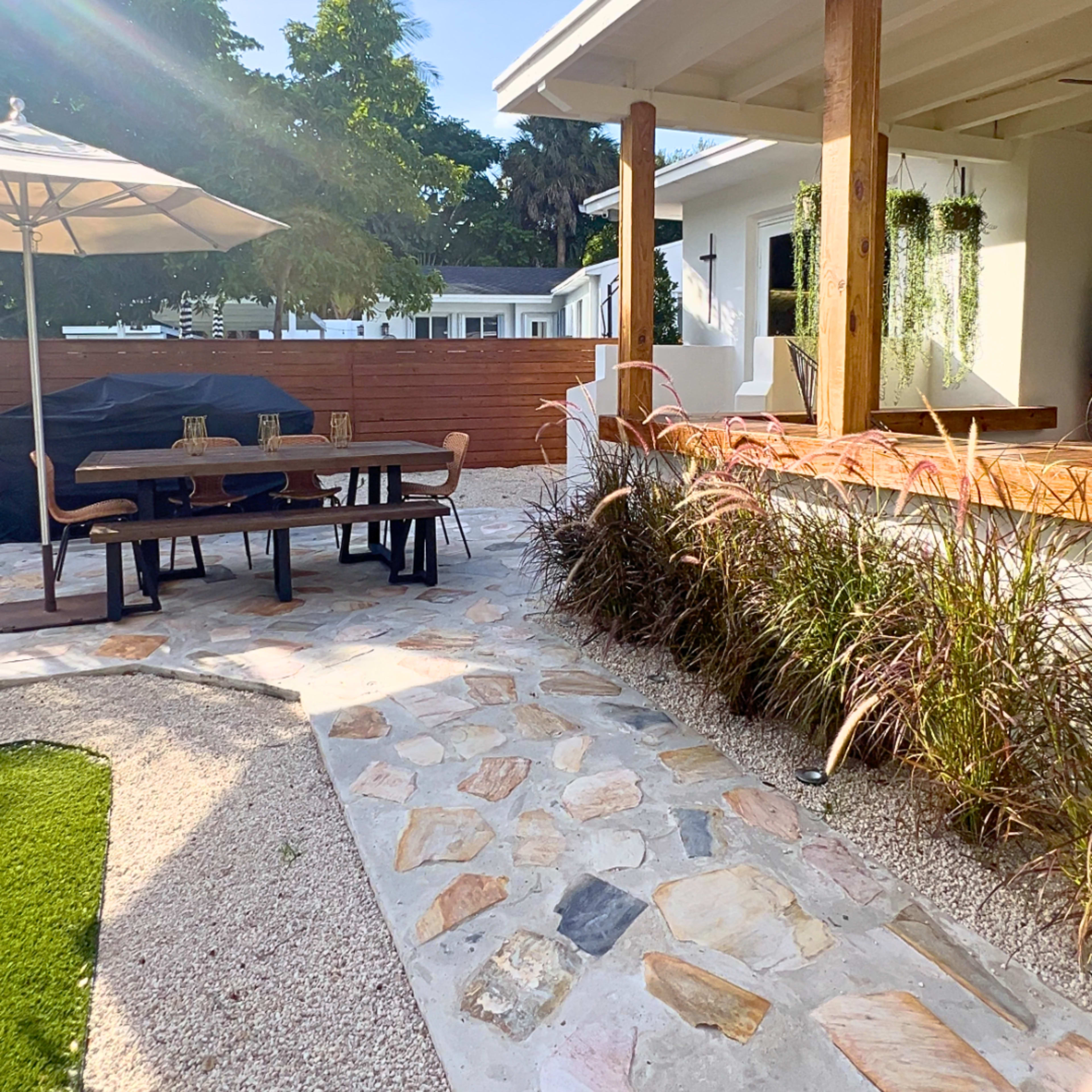 A landscaped outdoor area featuring a stone pathway, a dining table with chairs, an umbrella, and a porch with wooden posts.