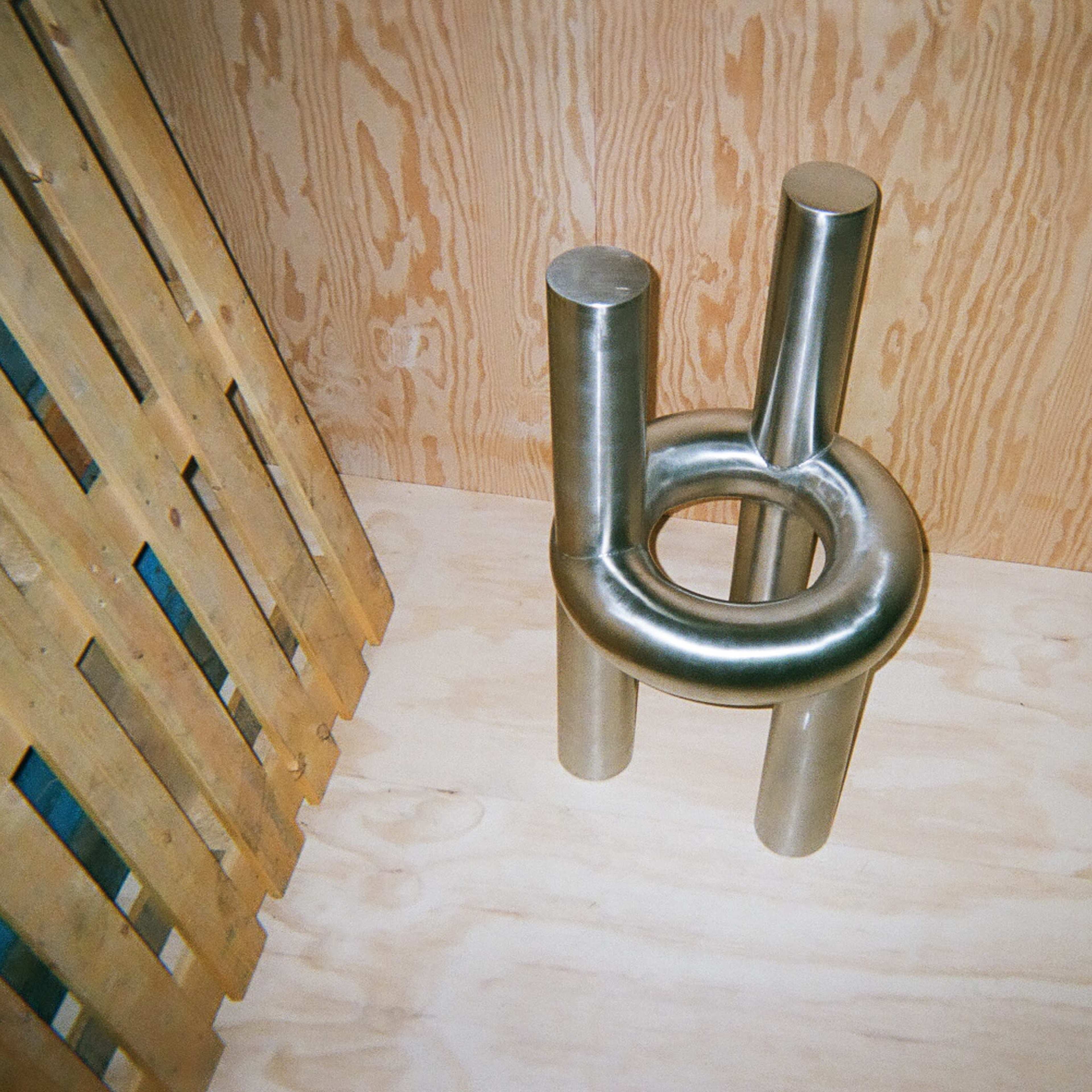 A metallic chair with a circular seat and two upright cylindrical legs placed on a wooden surface.