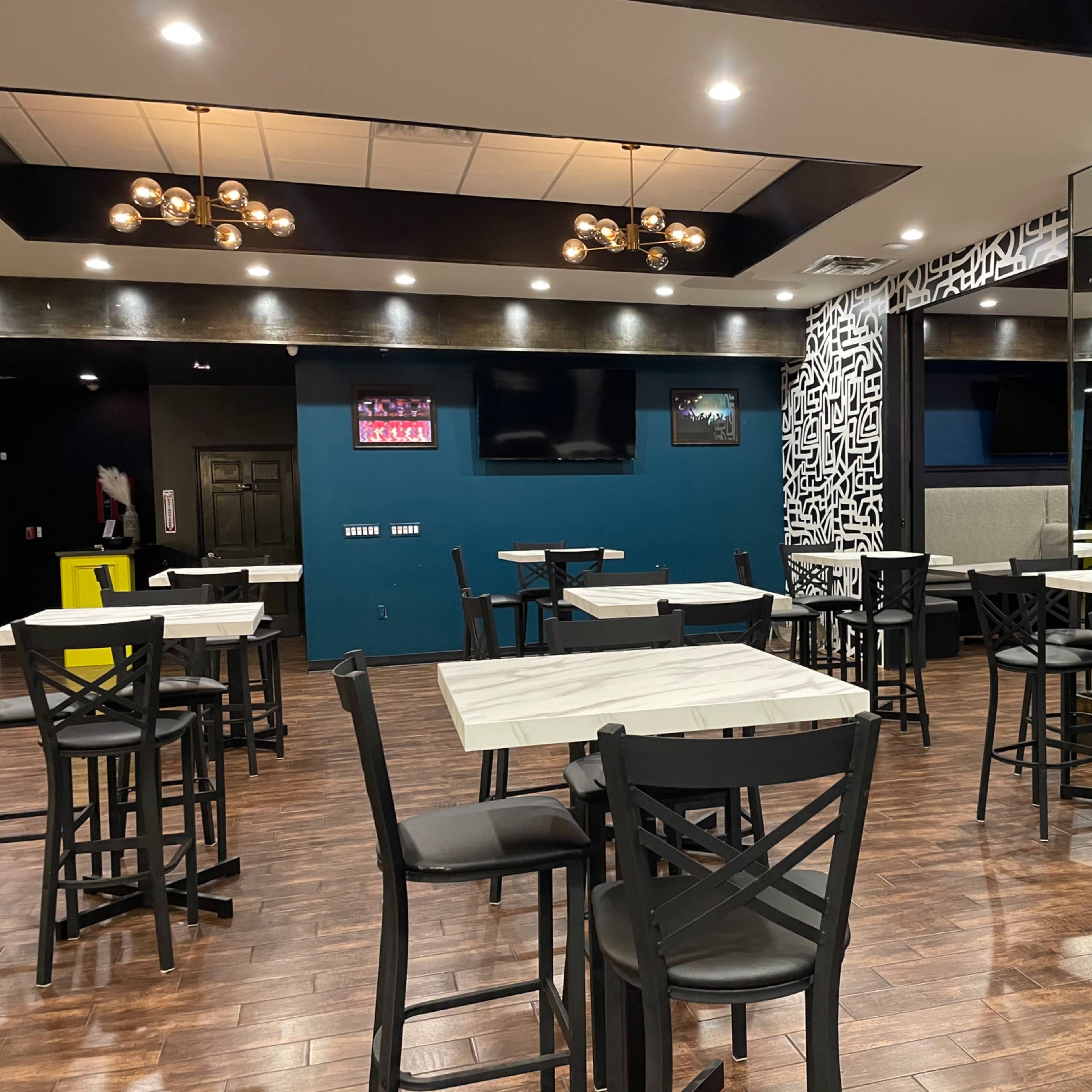 The photo shows a modern restaurant interior with vacant tables and chairs, dark wood flooring, and an accent wall featuring a geometric design.