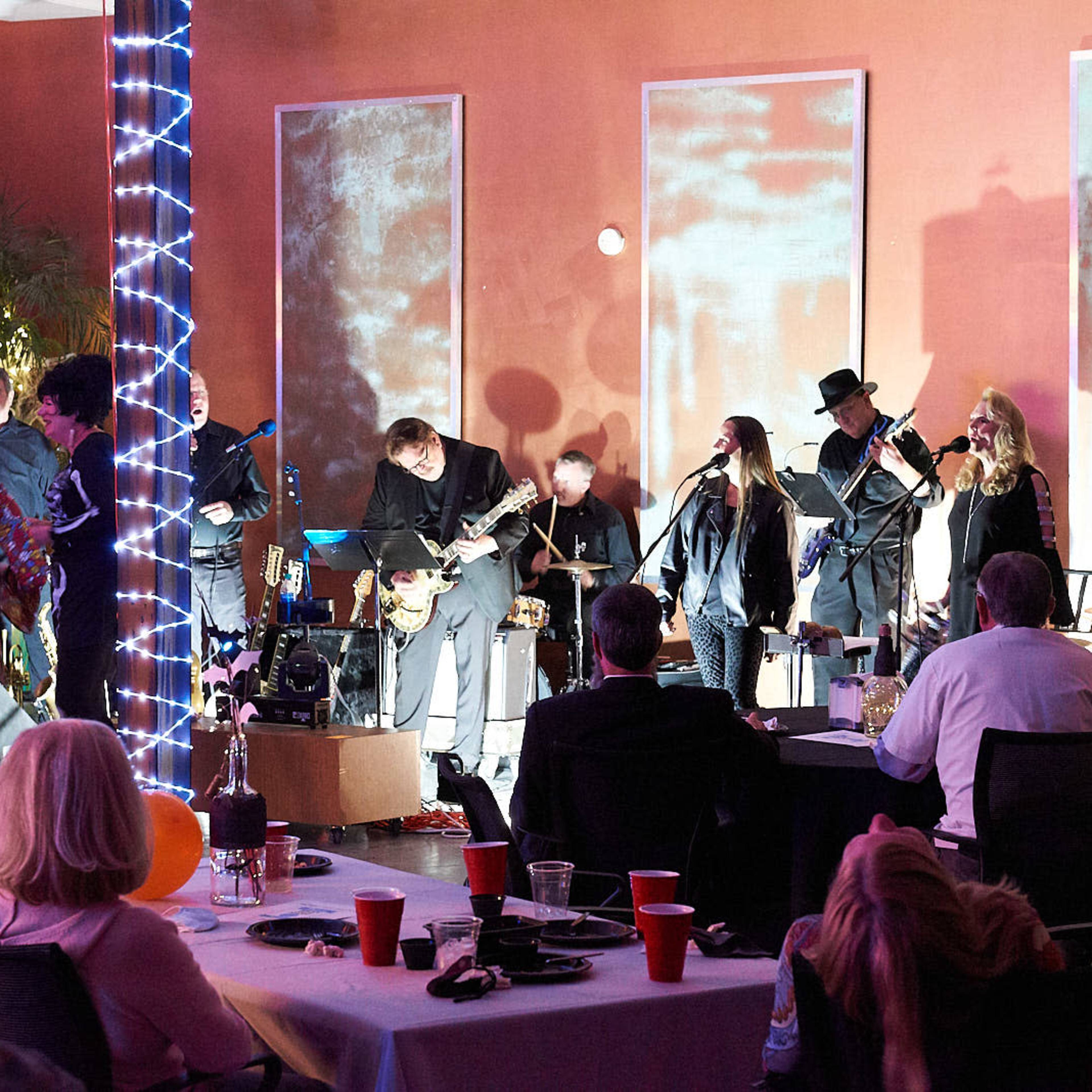 A live band performs on stage in a decorated venue with scattered tables and attendees enjoying the event.