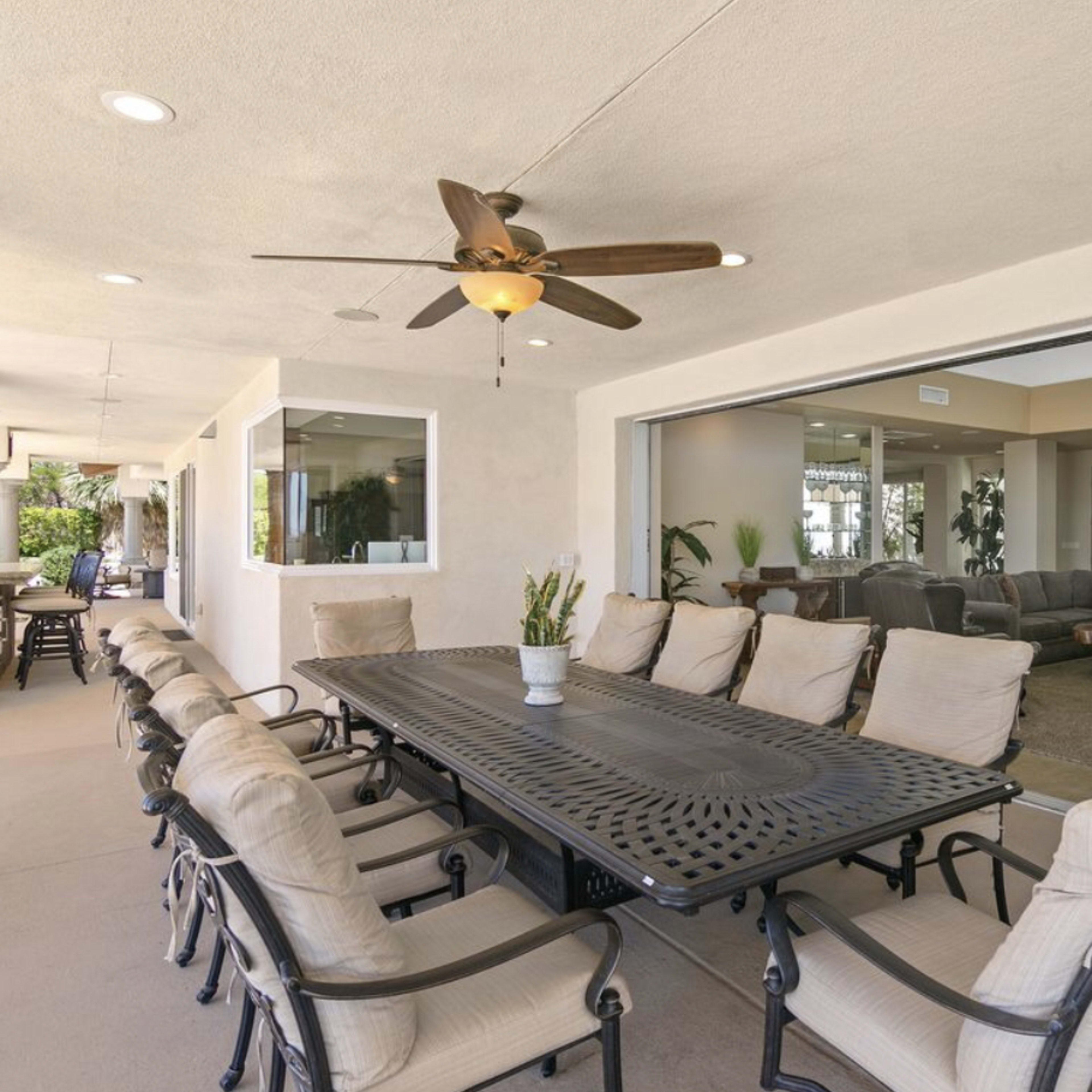 A covered patio area featuring a large dining table surrounded by chairs, with a ceiling fan above and a view of an interior living space through large glass windows.