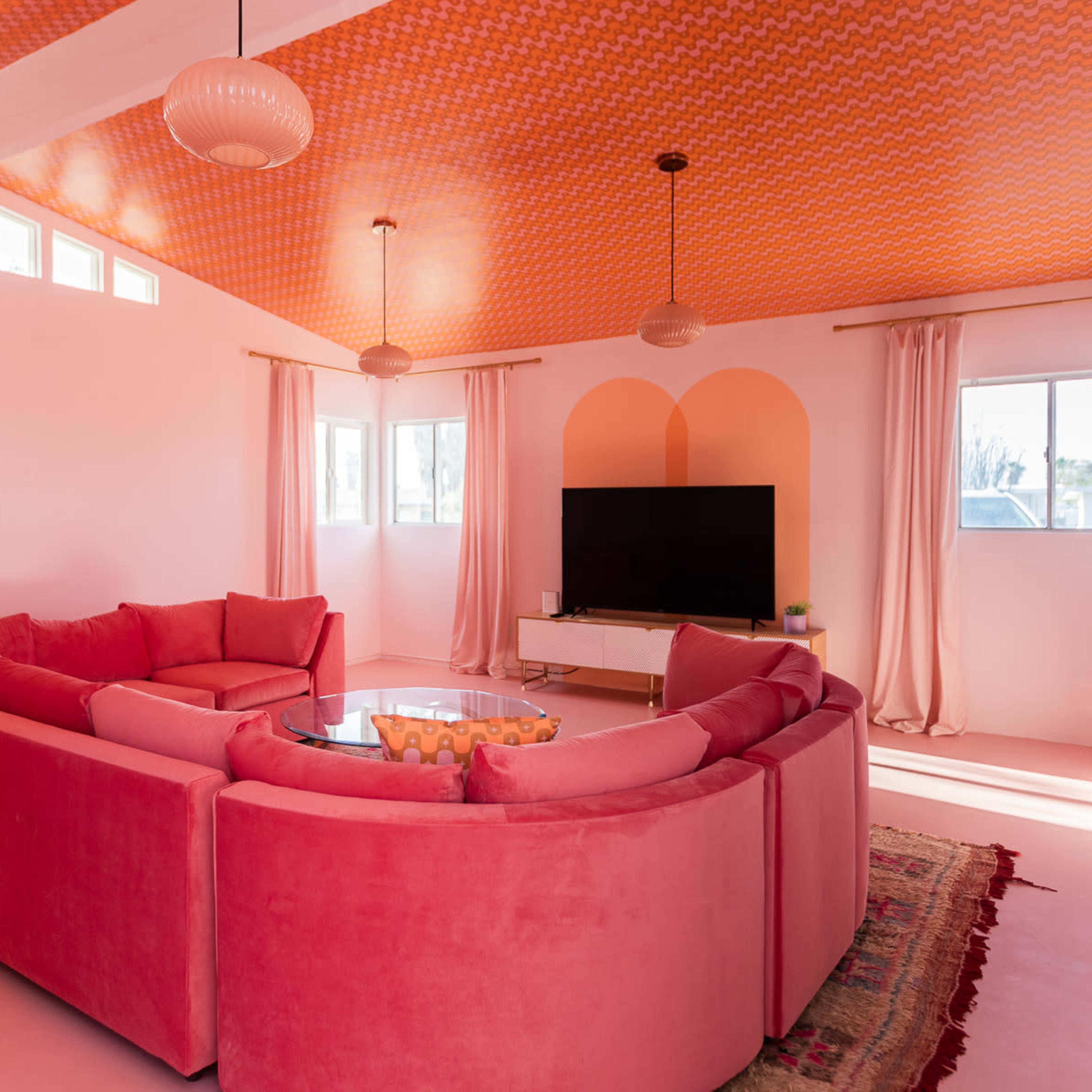 The room features a large, circular pink sofa arranged around a glass coffee table, with a television on a white stand against a wall and large windows providing natural light.