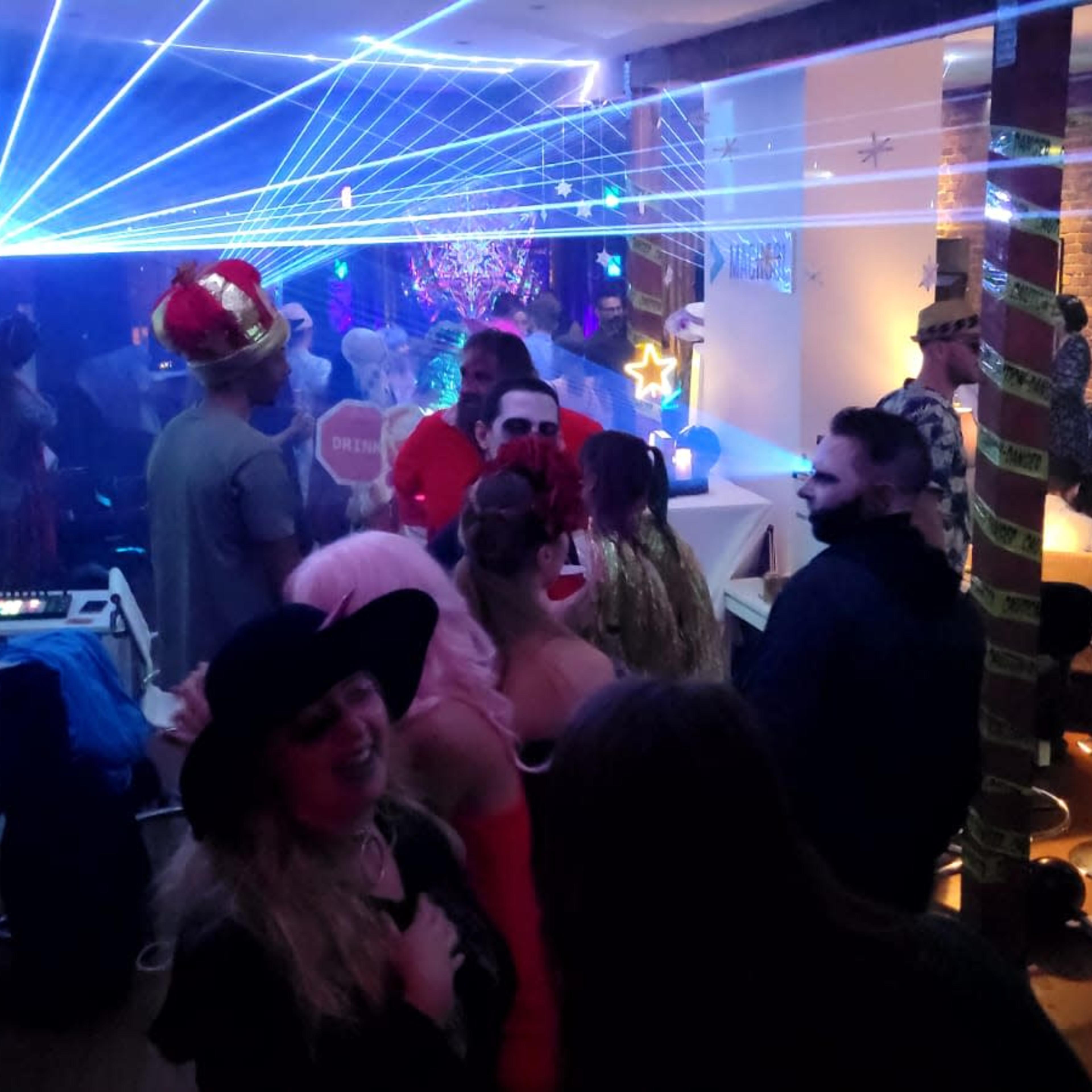 A lively party scene features guests mingling under colorful laser lights and decorations in a dimly lit room.