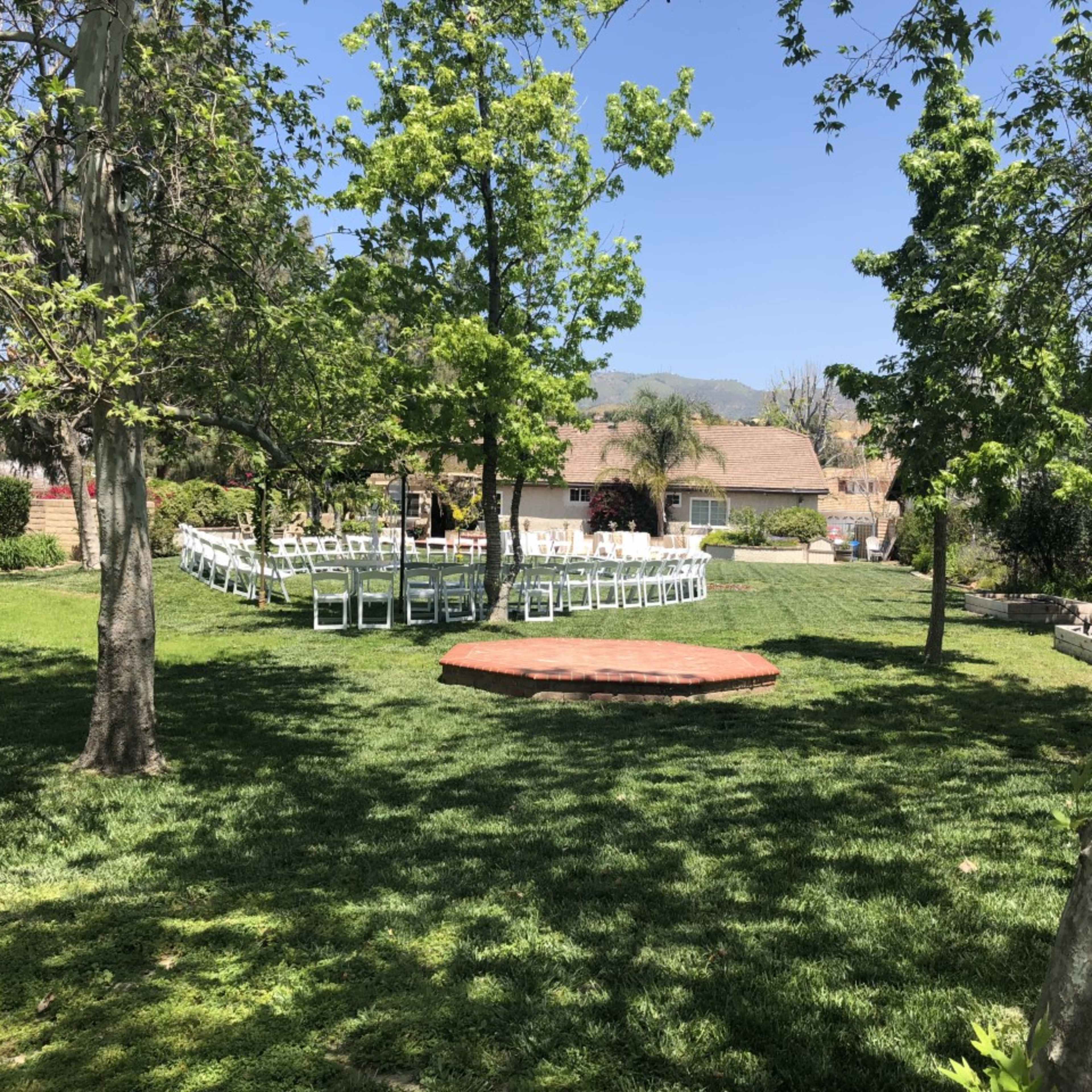 A landscaped outdoor area features a stage and rows of white chairs set up for an event surrounded by trees and green grass.