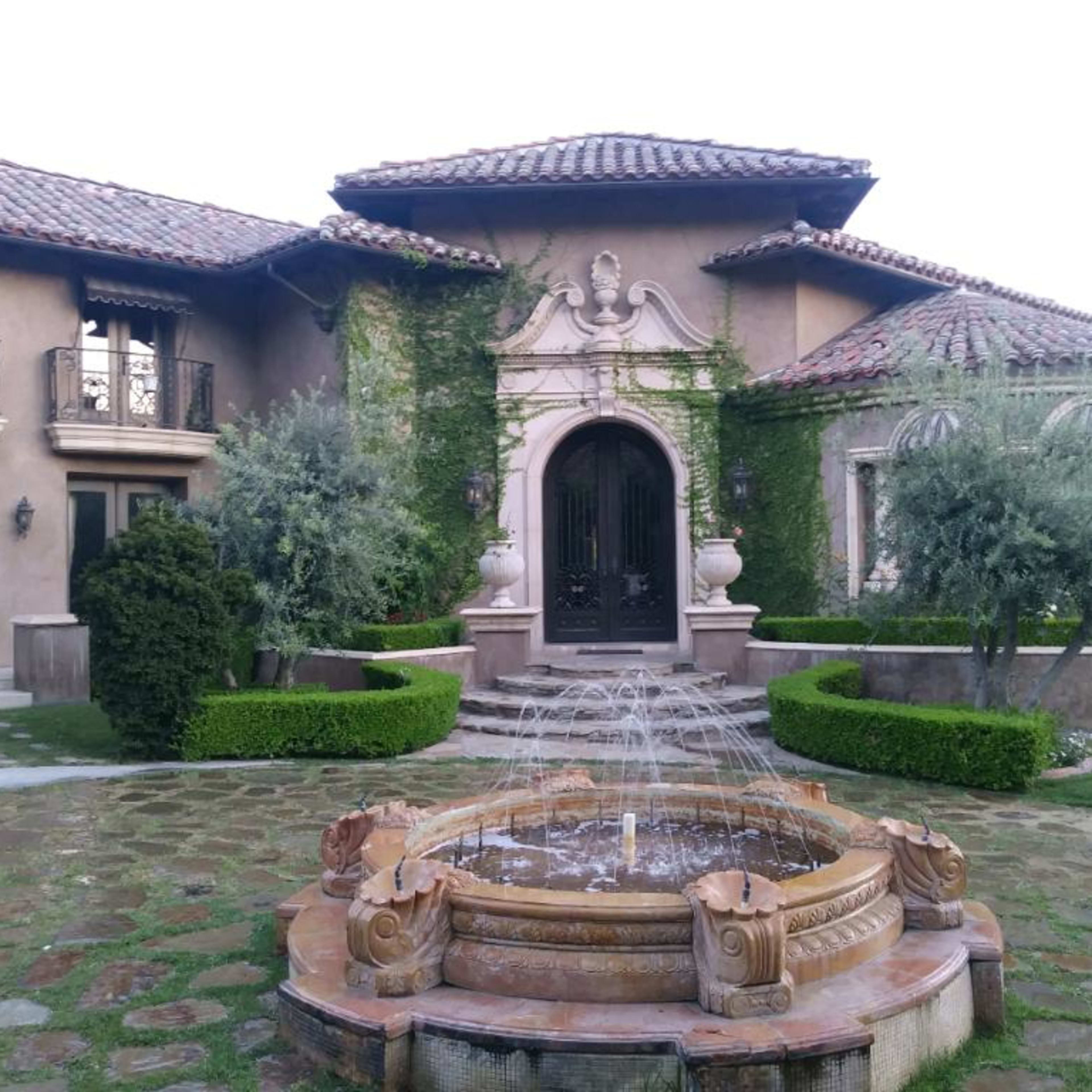 A luxurious home with a grand entrance, surrounded by manicured landscaping and a central fountain.