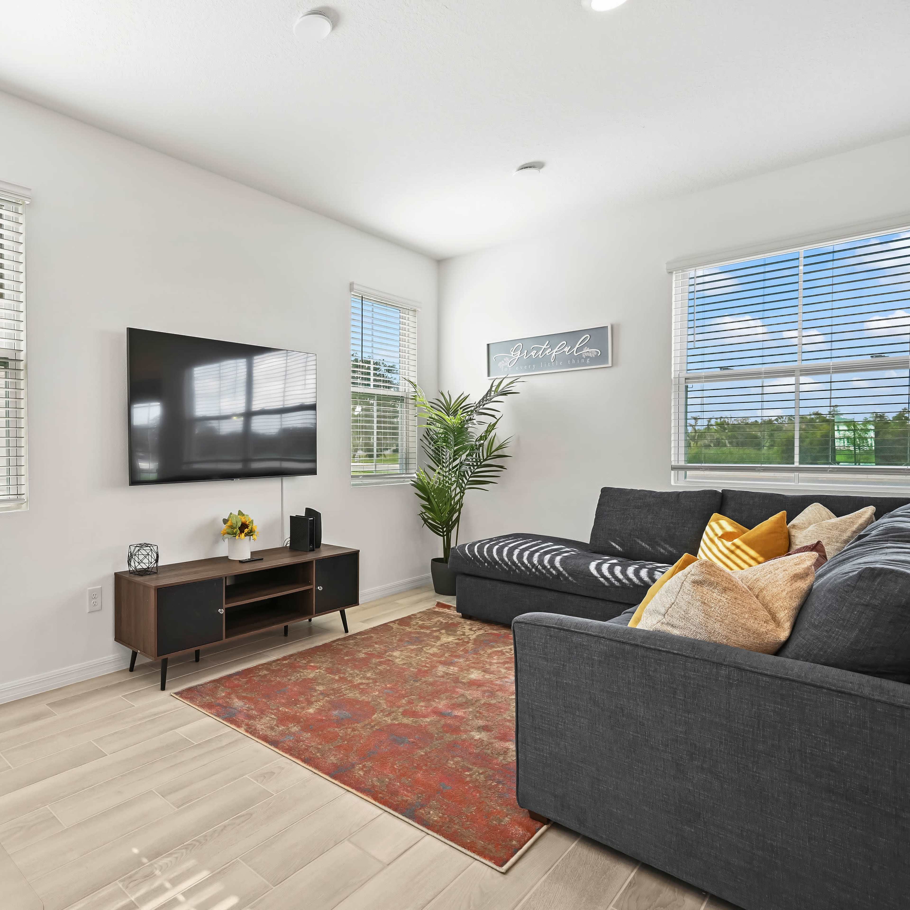 A modern living room features a sectional sofa, a wall-mounted television, a desk with a lamp, and a decorative rug.