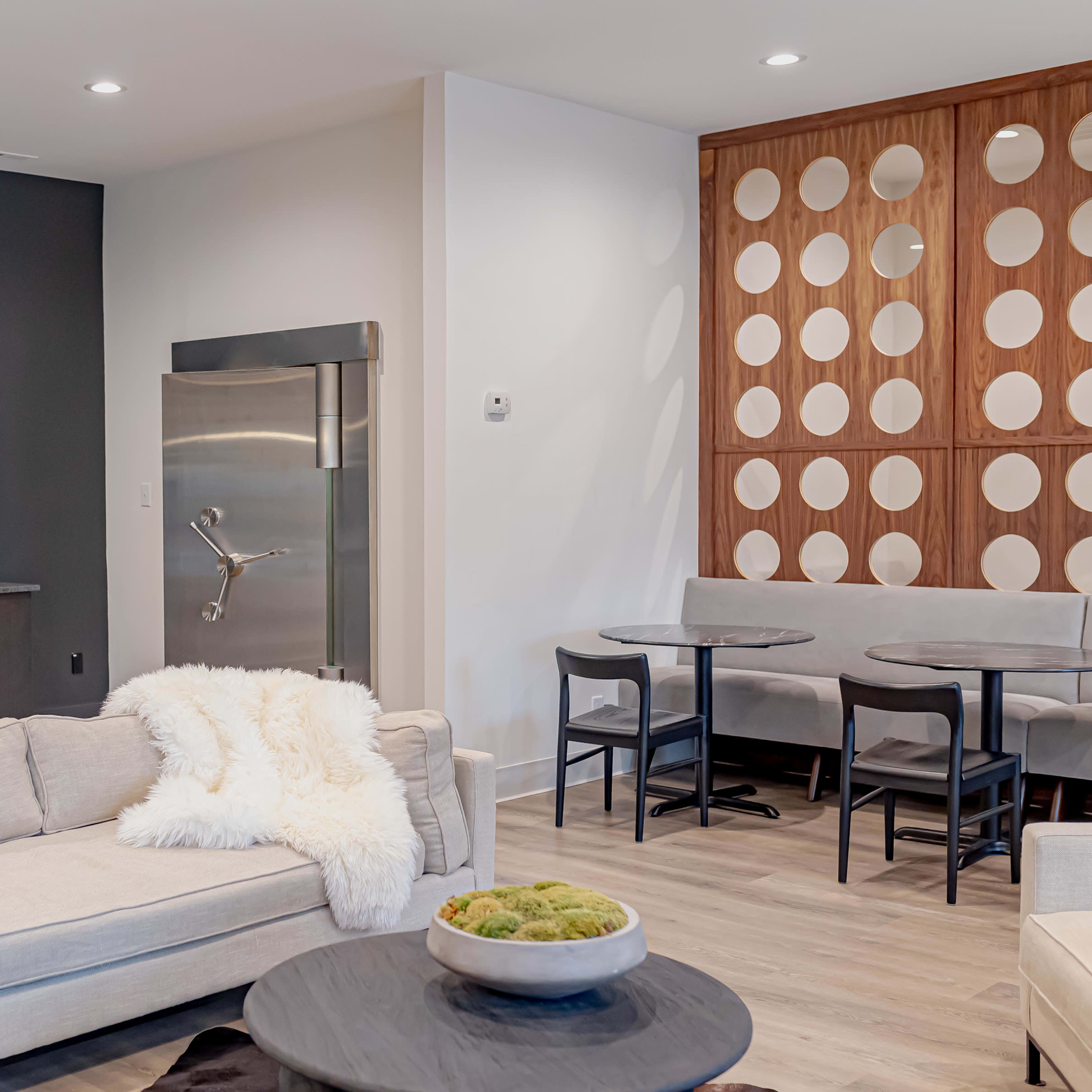 A modern living space featuring a light-colored sofa, a round black coffee table, a kitchen area with stainless steel appliances, and a dining area with round tables and minimalist seating.