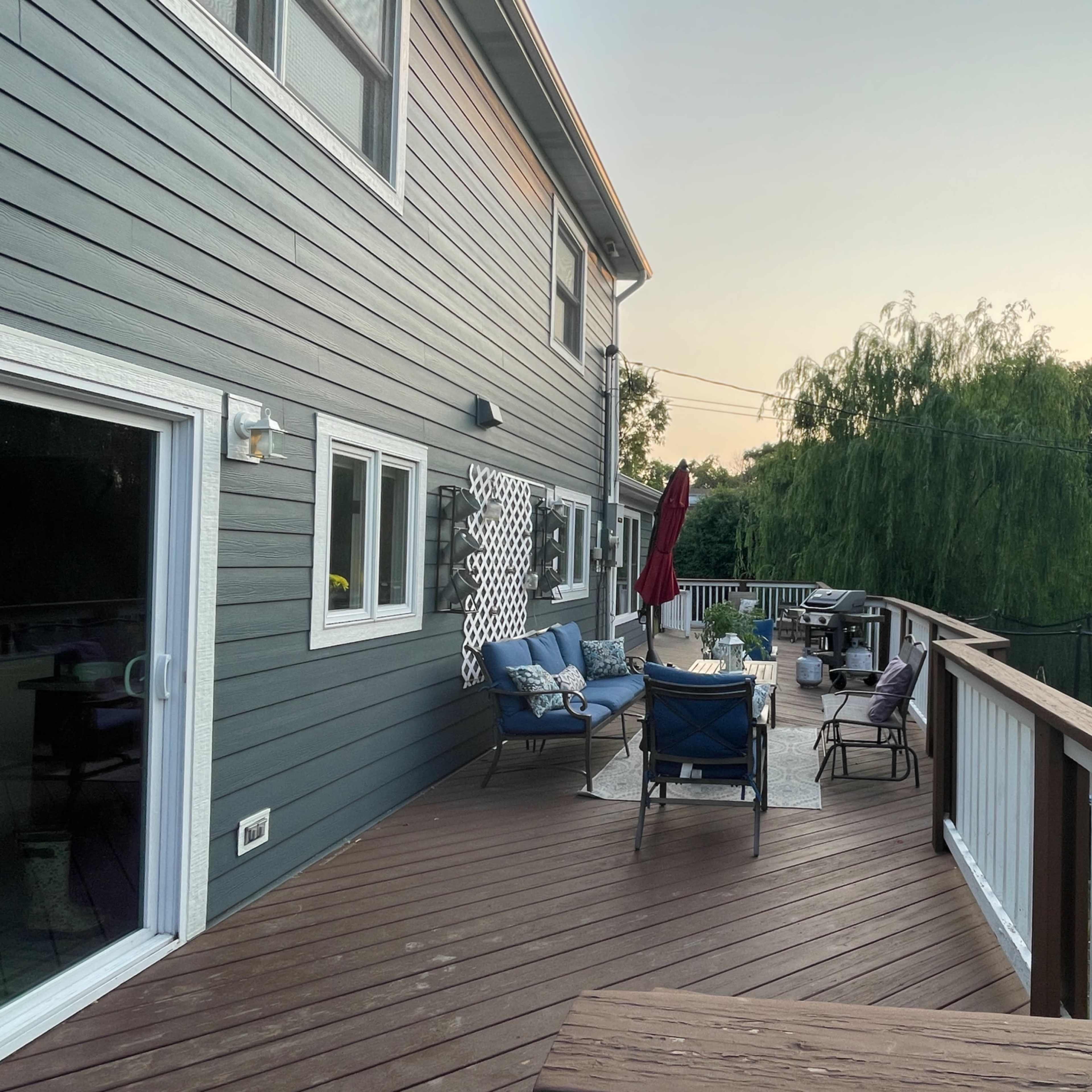 A wooden deck wraps around a house, featuring outdoor furniture and a red umbrella, with trees and a trampoline visible in the background.