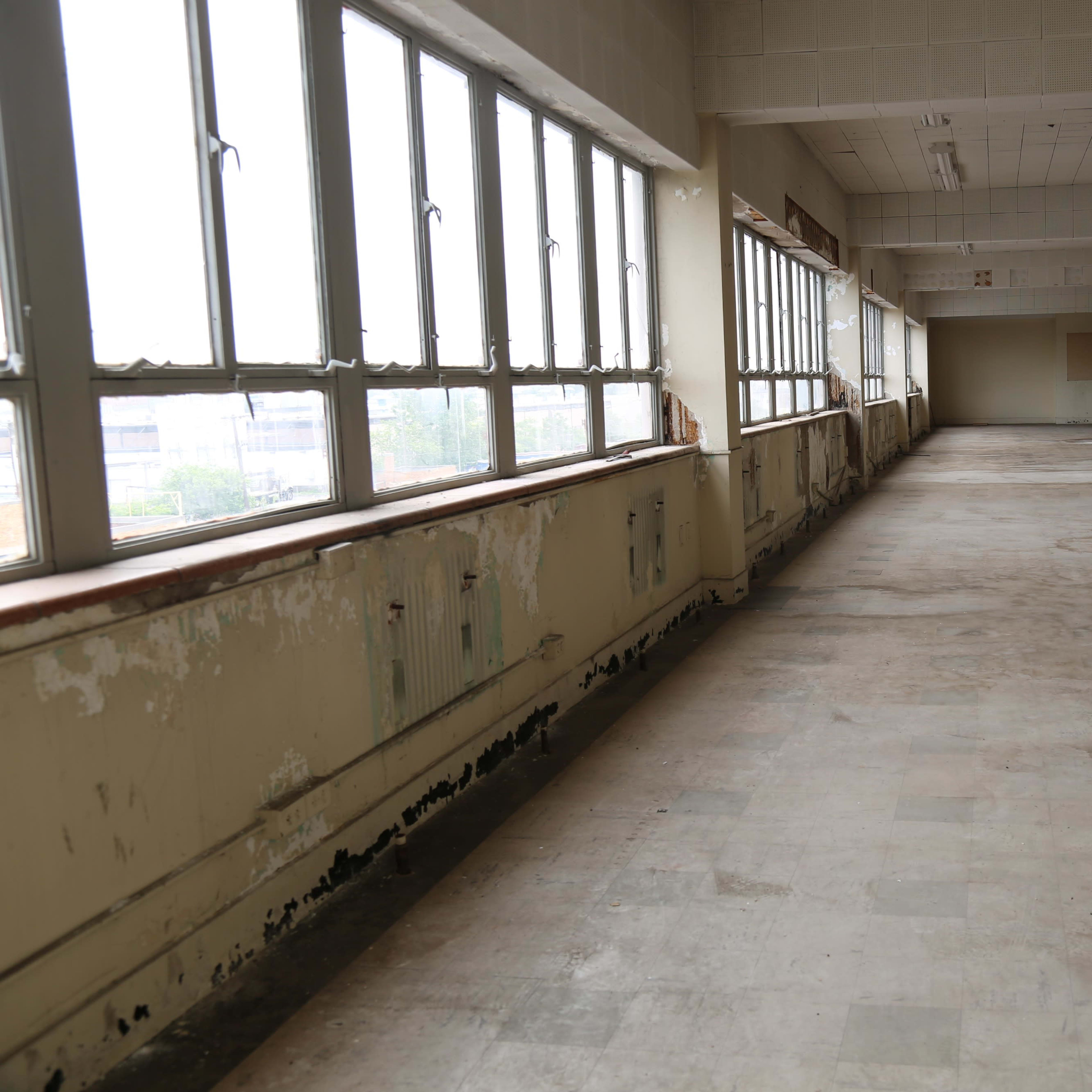 A large, empty room with worn flooring and windows along one side, allowing natural light to enter.
