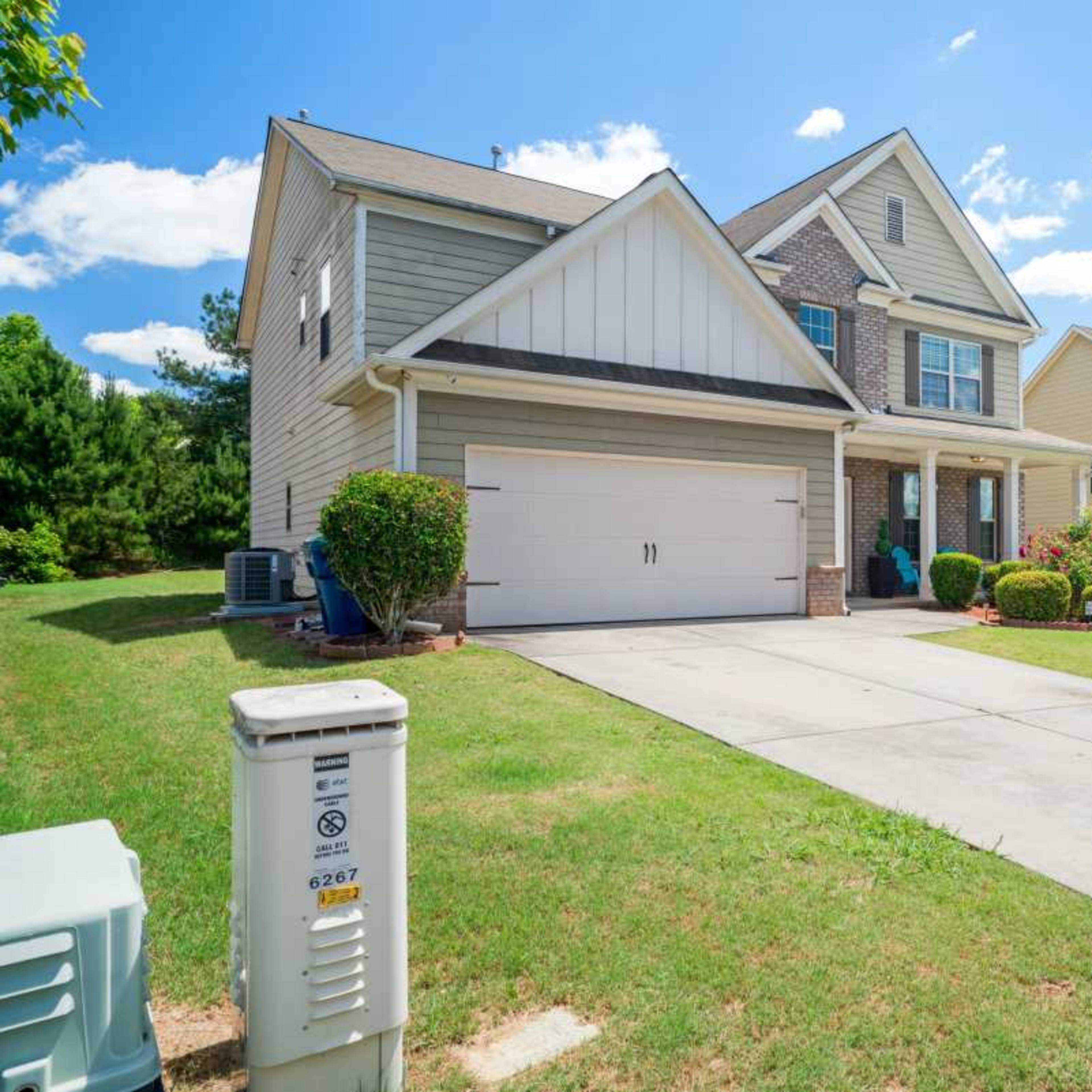 A two-story house with a front porch and a double garage is situated on a green lawn with nearby trees and shrubs.