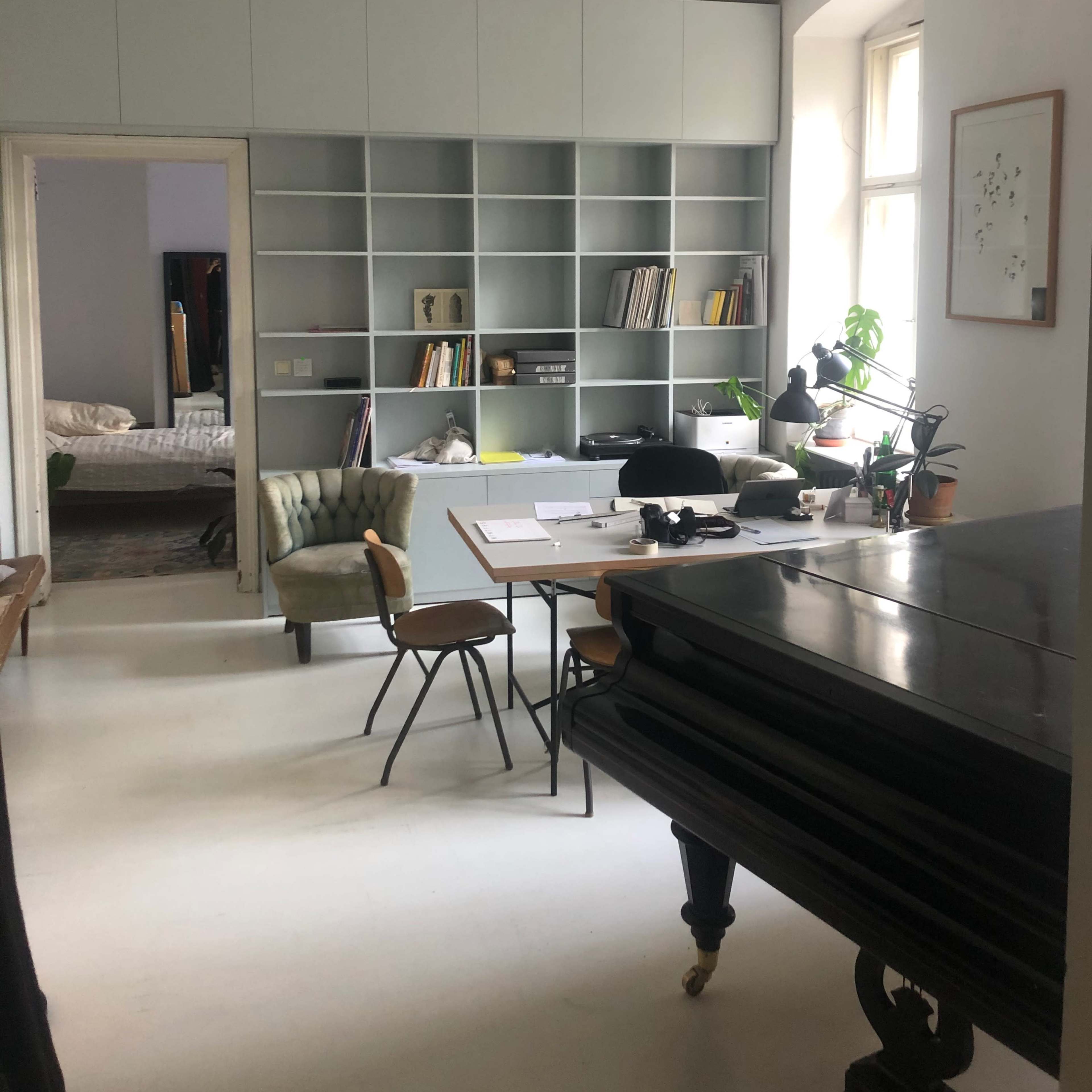 A modern room with a desk, chair, piano, and built-in shelves filled with books, located next to a mirror and a bedroom.