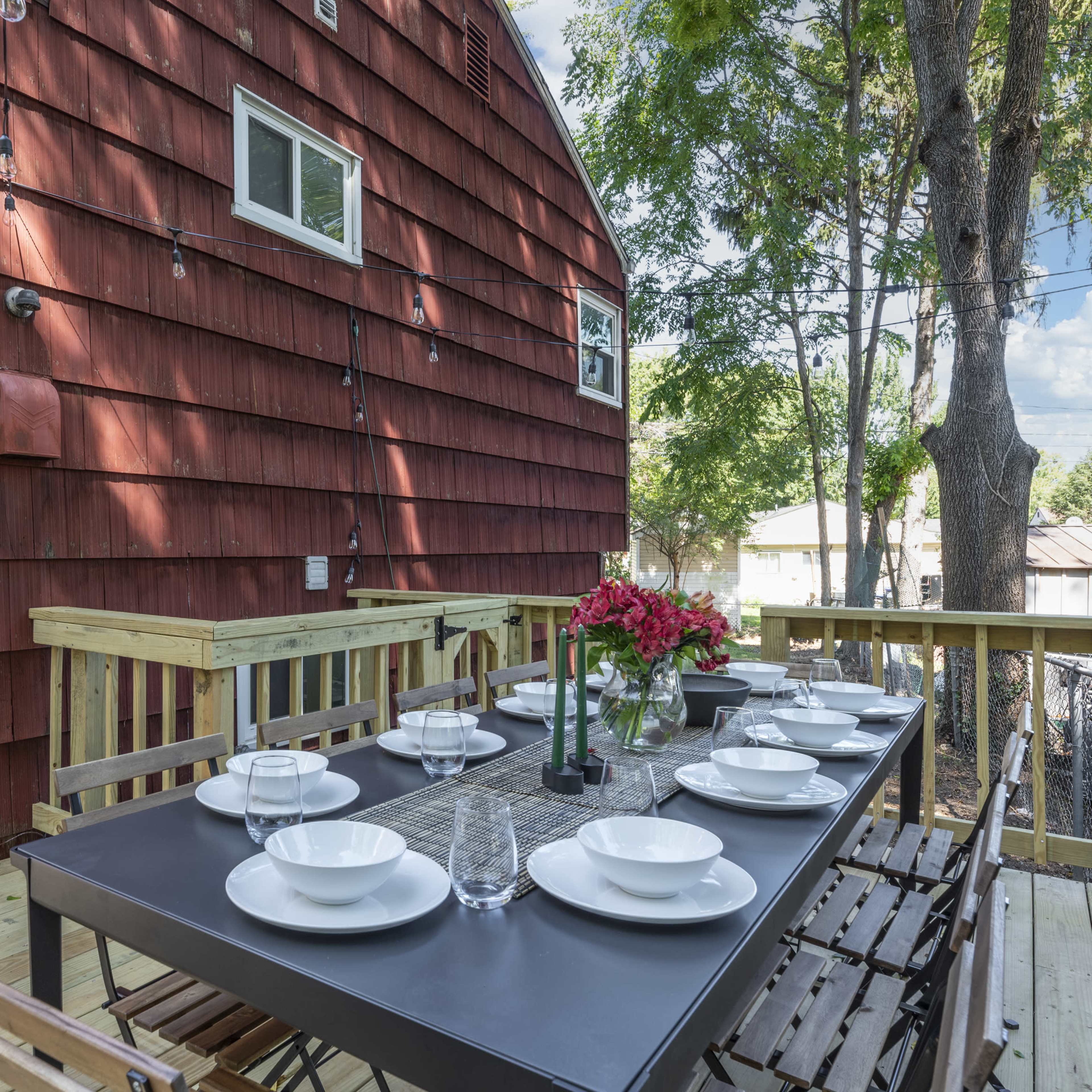 A wooden deck with a long dining table set for eight, overlooks a red-shingled house and surrounding trees.