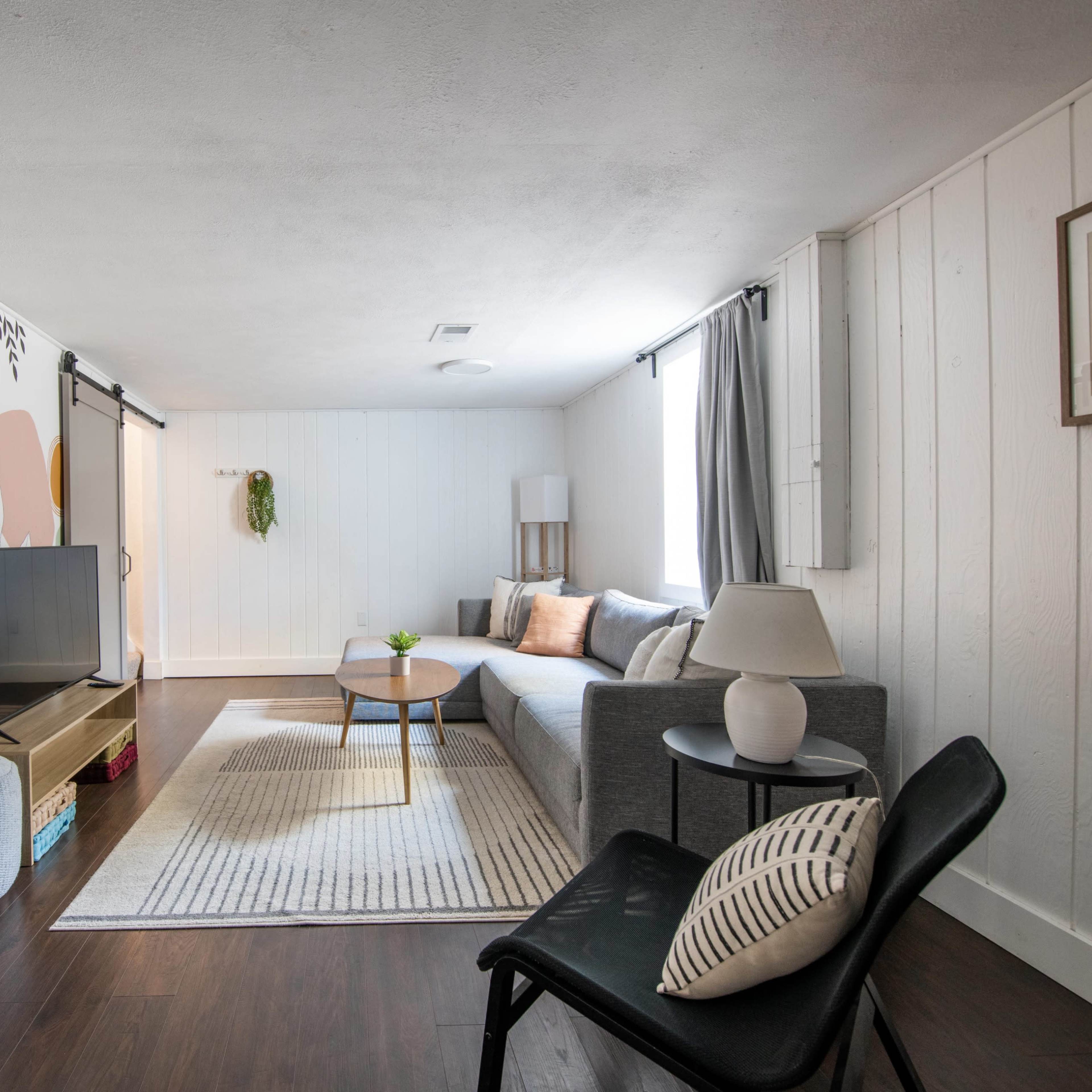 A modern living room with a gray sofa, a television on a light wooden stand, a coffee table, and a decorative wall featuring colorful shapes.