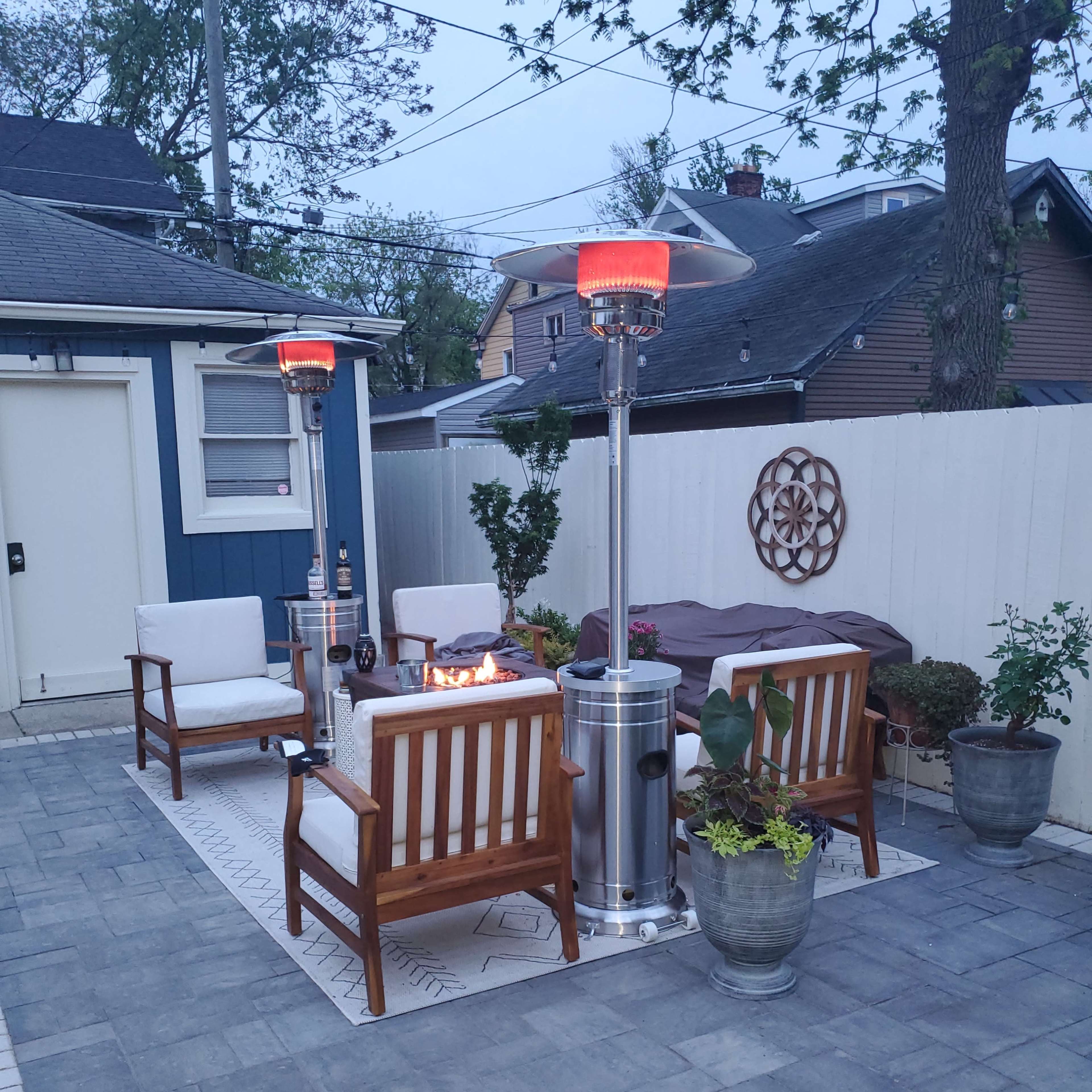 A cozy outdoor seating area features two patio heaters, four wooden chairs, a fire pit, and potted plants on a stone patio.
