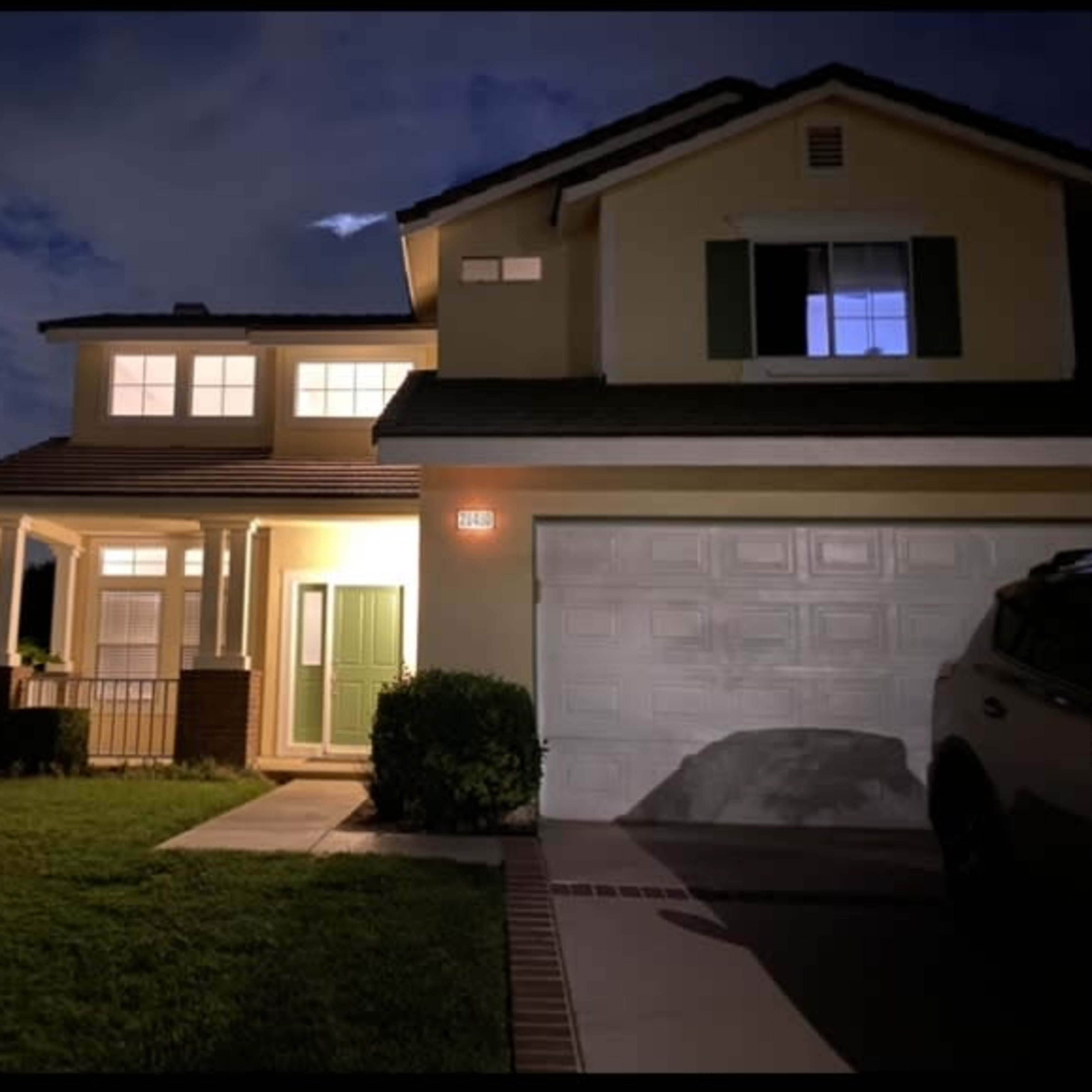 A two-story residential house is illuminated by a porch light, with a neatly maintained lawn and a vehicle parked in the driveway at night.