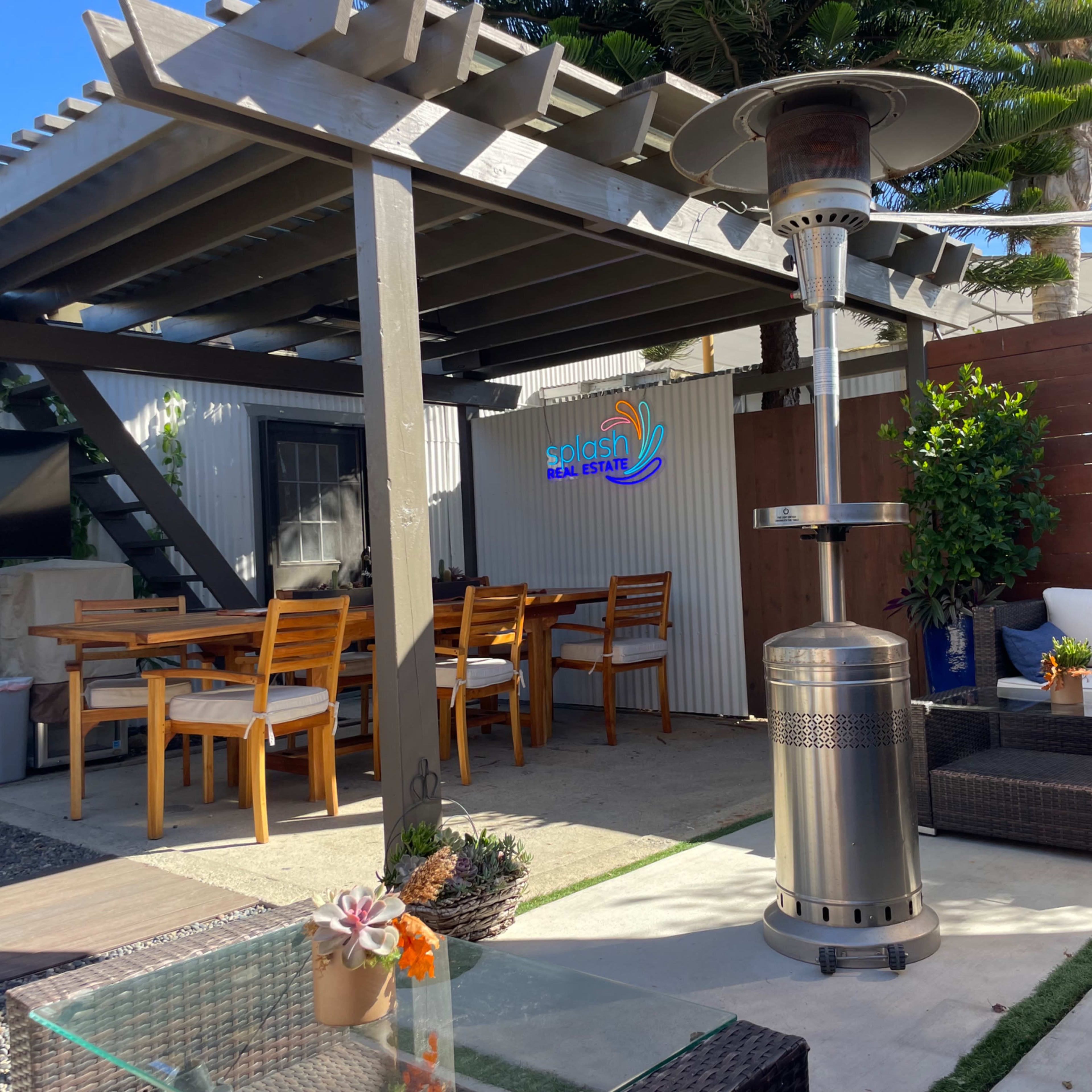 A patio area features a dining table, comfortable seating, and a patio heater under a pergola surrounded by greenery.