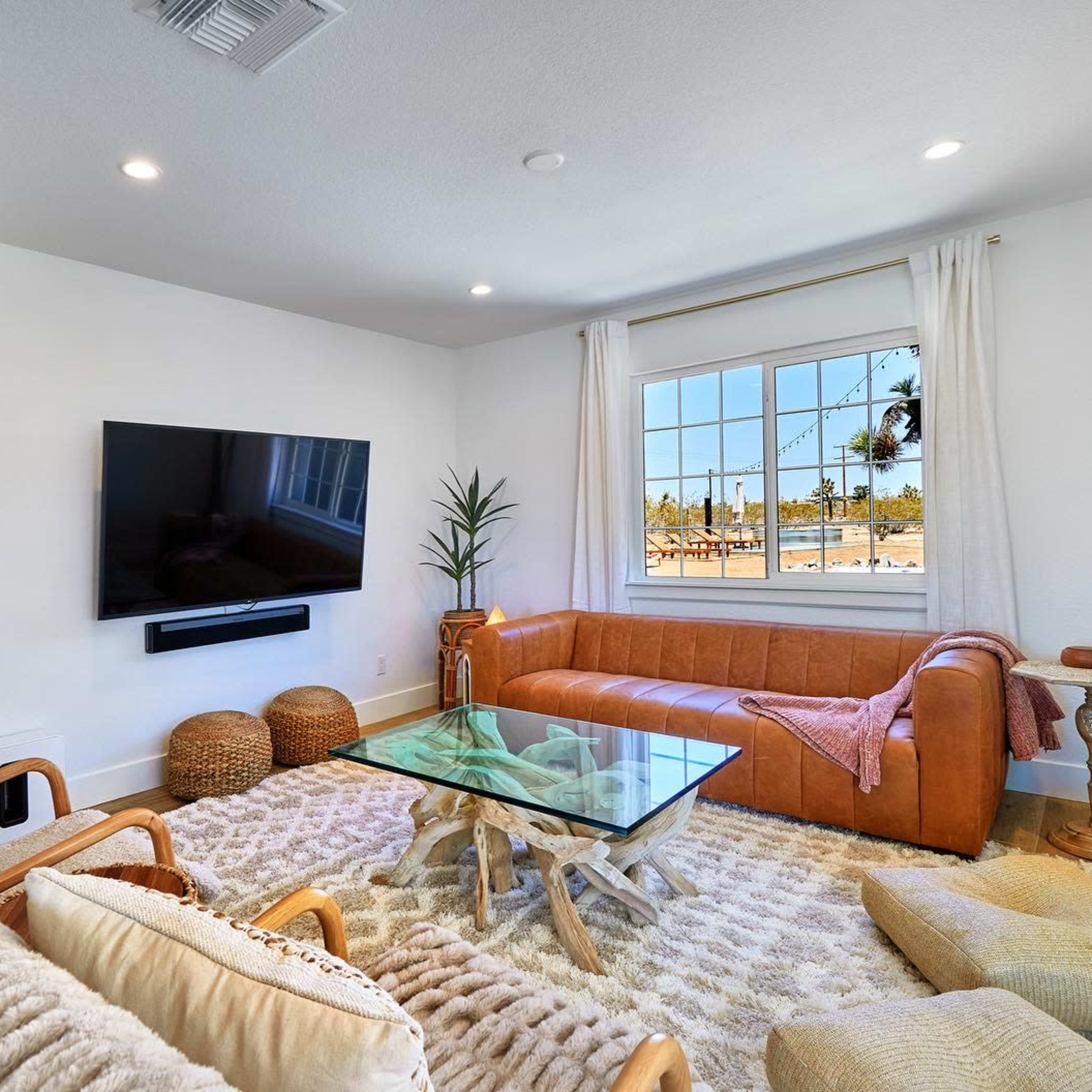 A modern living room features a large flat-screen TV mounted on the wall, a brown leather sofa, a glass coffee table, and a textured rug.