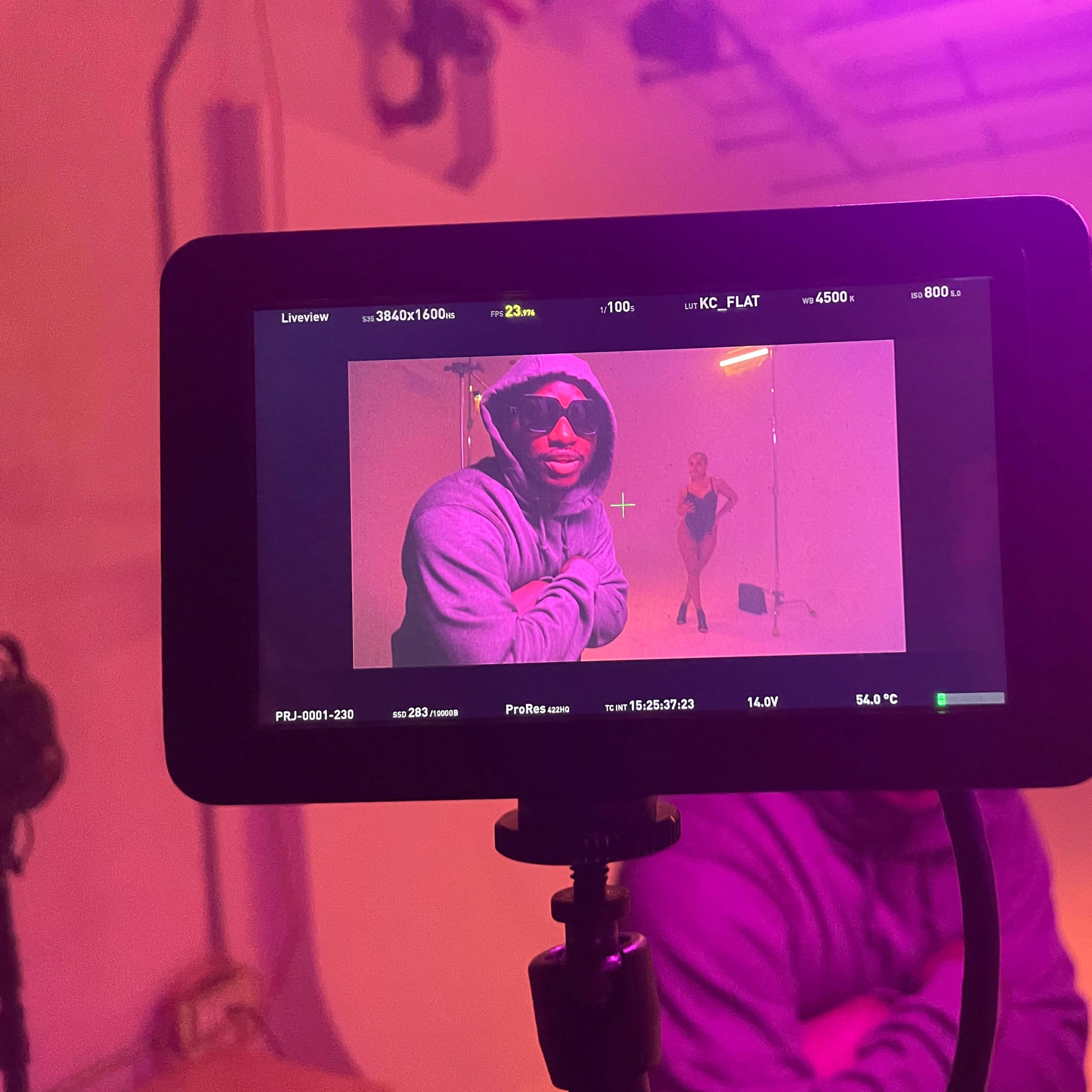A camera monitor displays a man posing in front of a woman in a dance outfit, illuminated by purple lighting in a studio setting.
