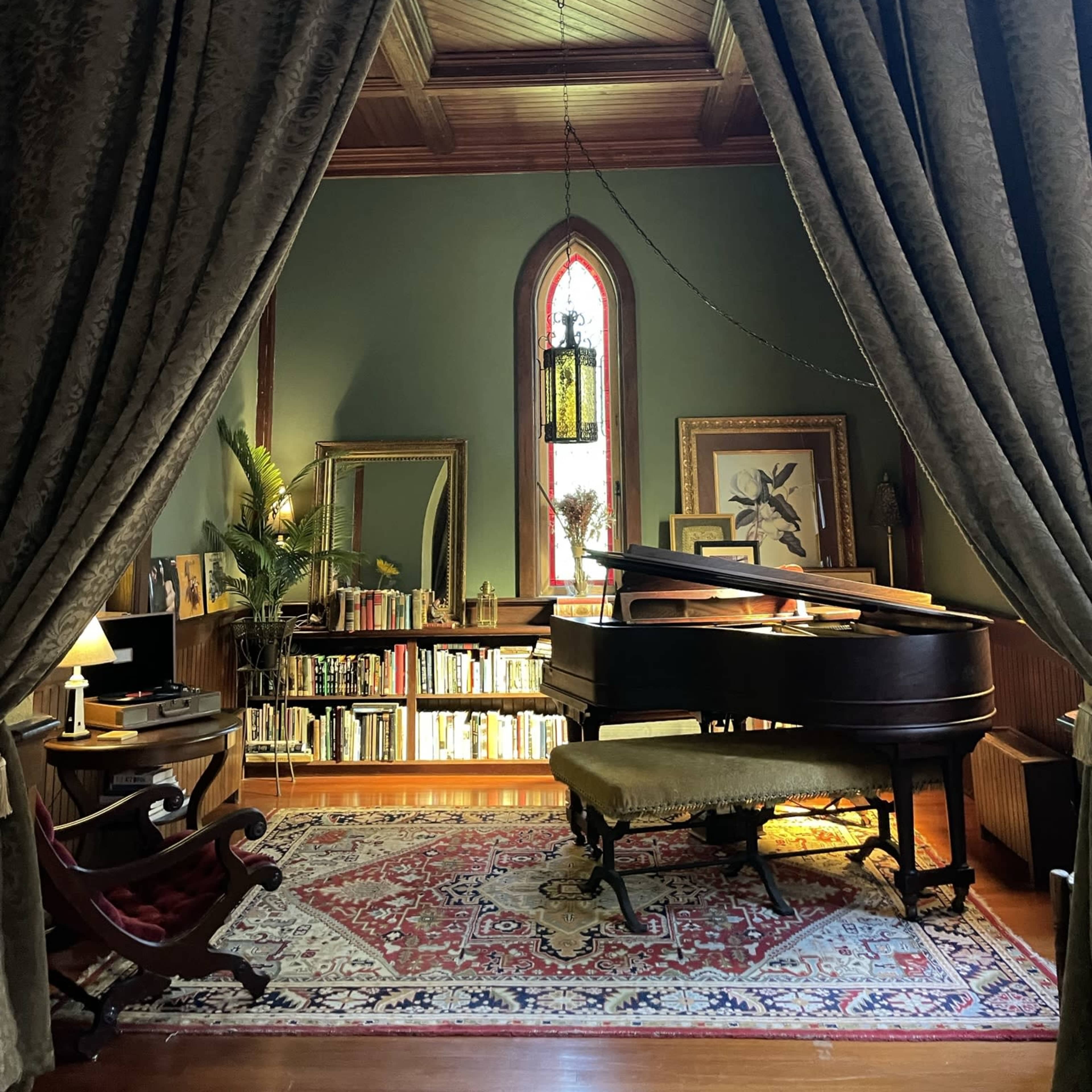 A grand piano is positioned in front of a window with stained glass, surrounded by bookshelves and a cozy seating area, framed by heavy curtains.