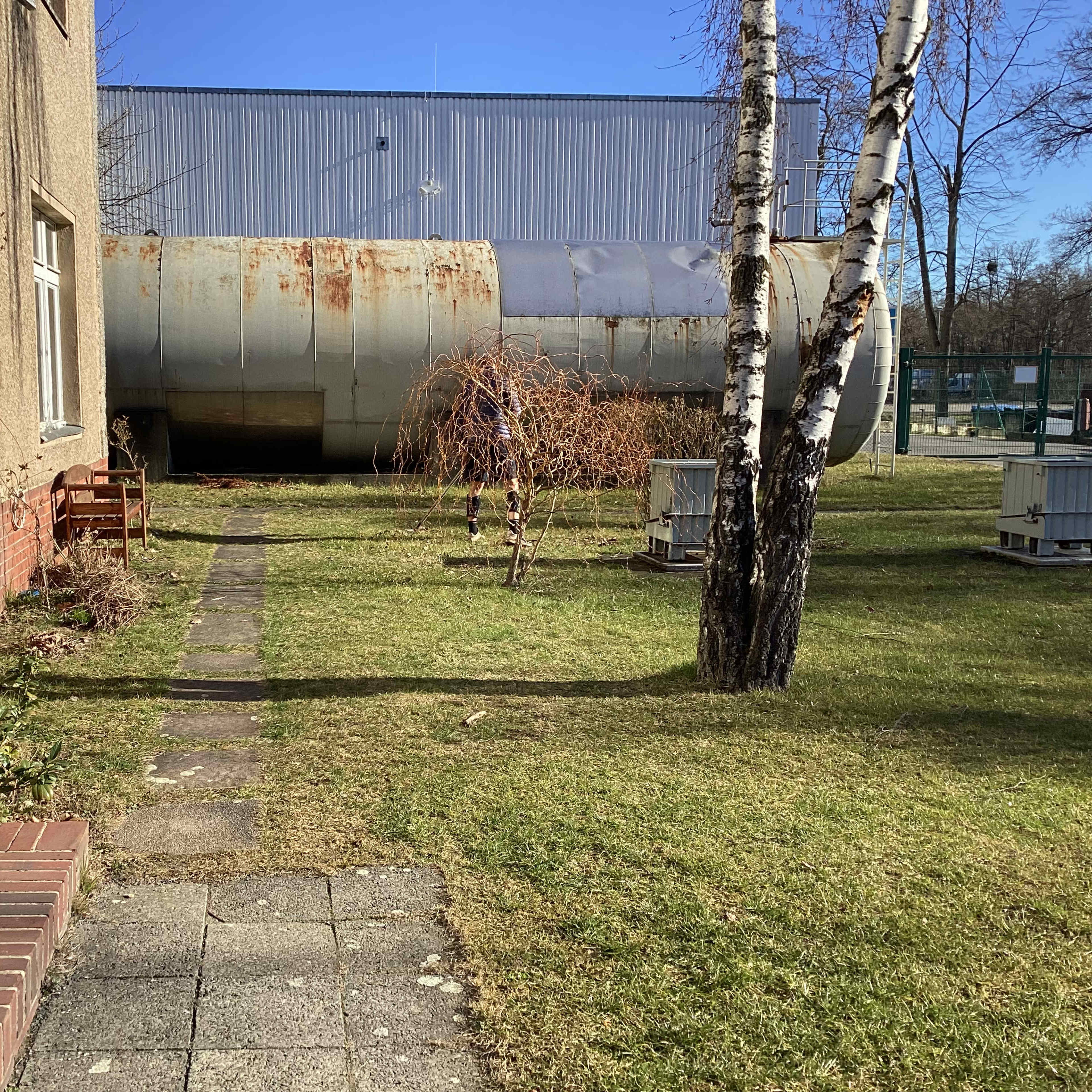 A pathway leading through a grassy area with trees and a large, rusted tank in the background near a building.