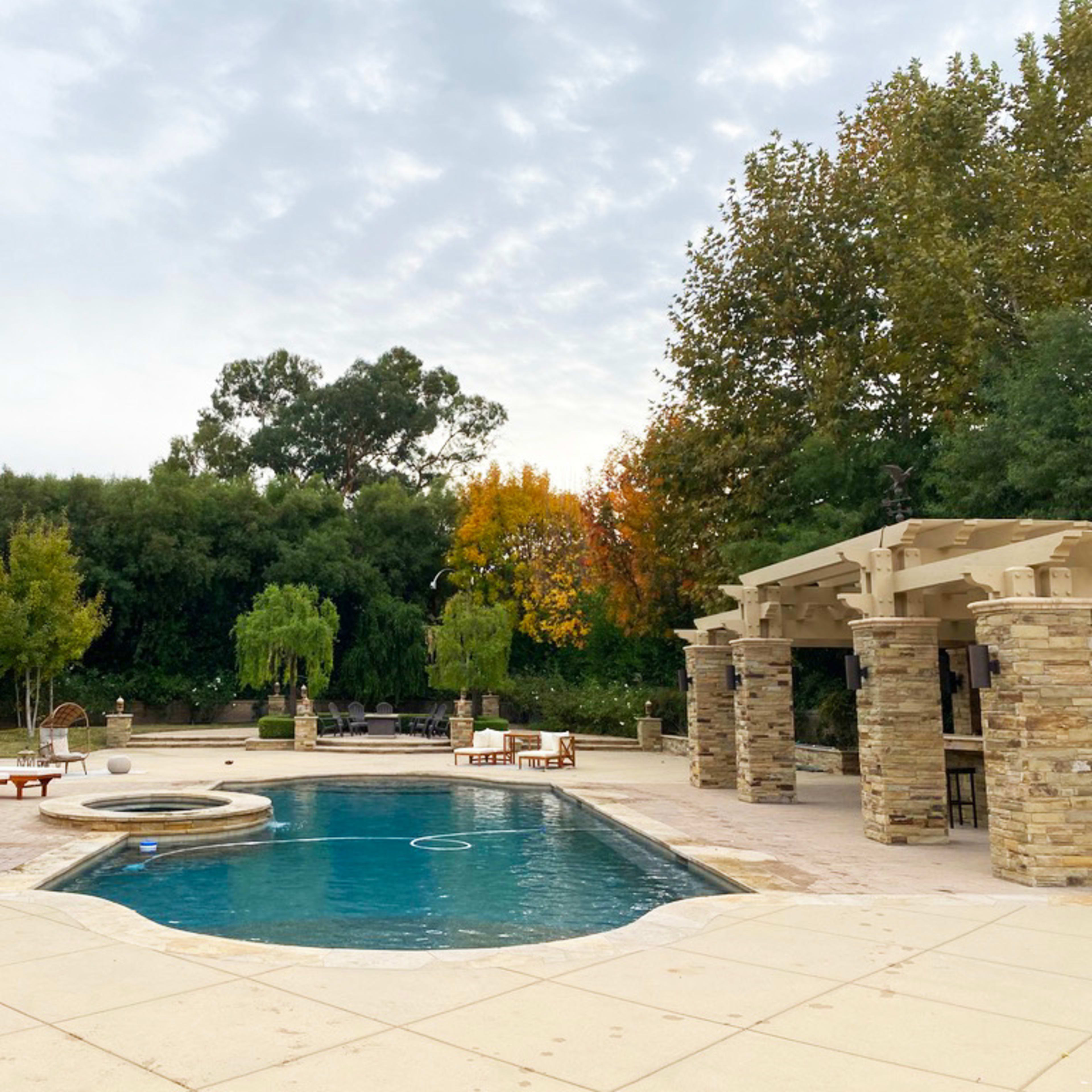 A backyard pool area surrounded by trees, lounge chairs, and a covered seating space.