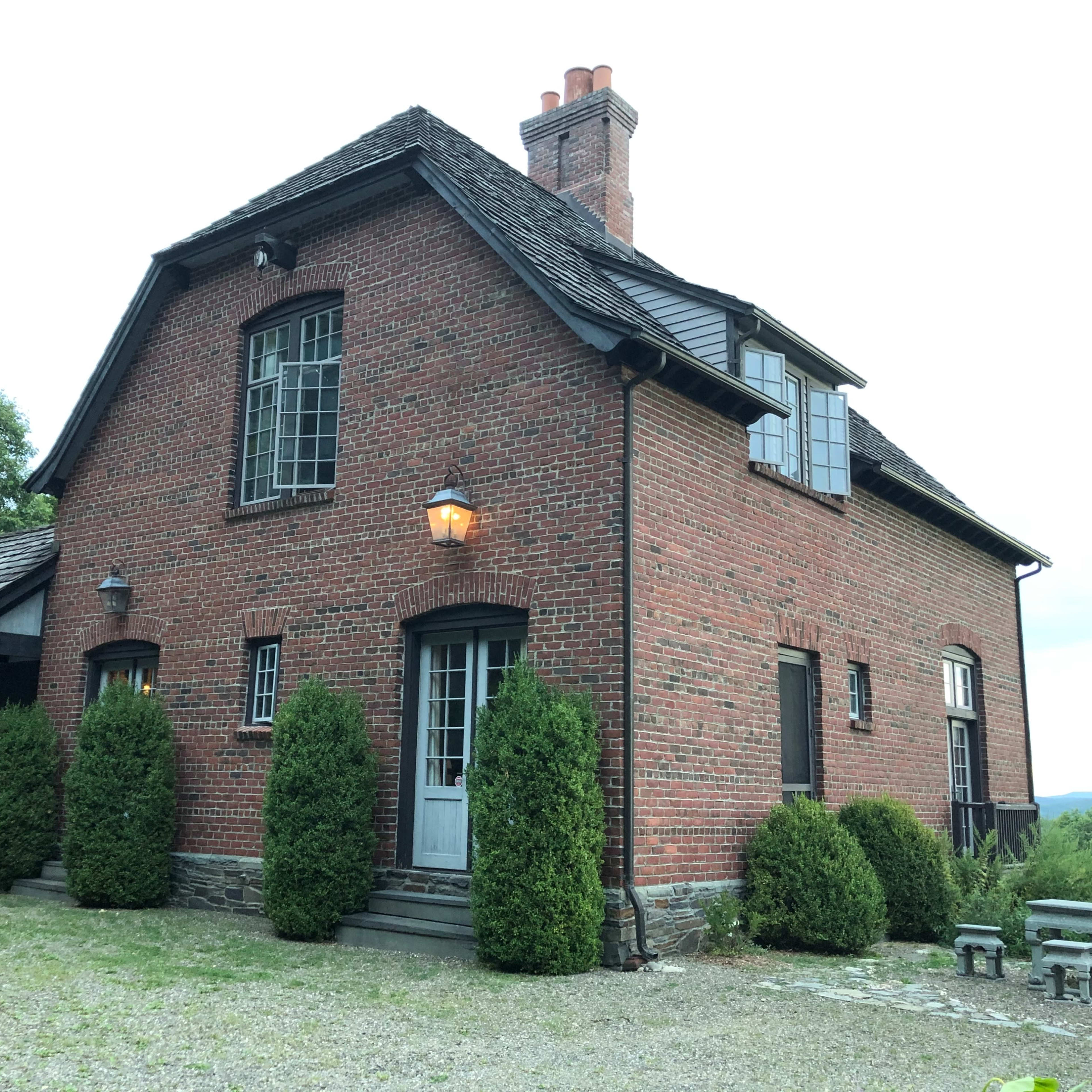 A two-story brick house with a chimney and several shrubs in front is situated on a gravel path.