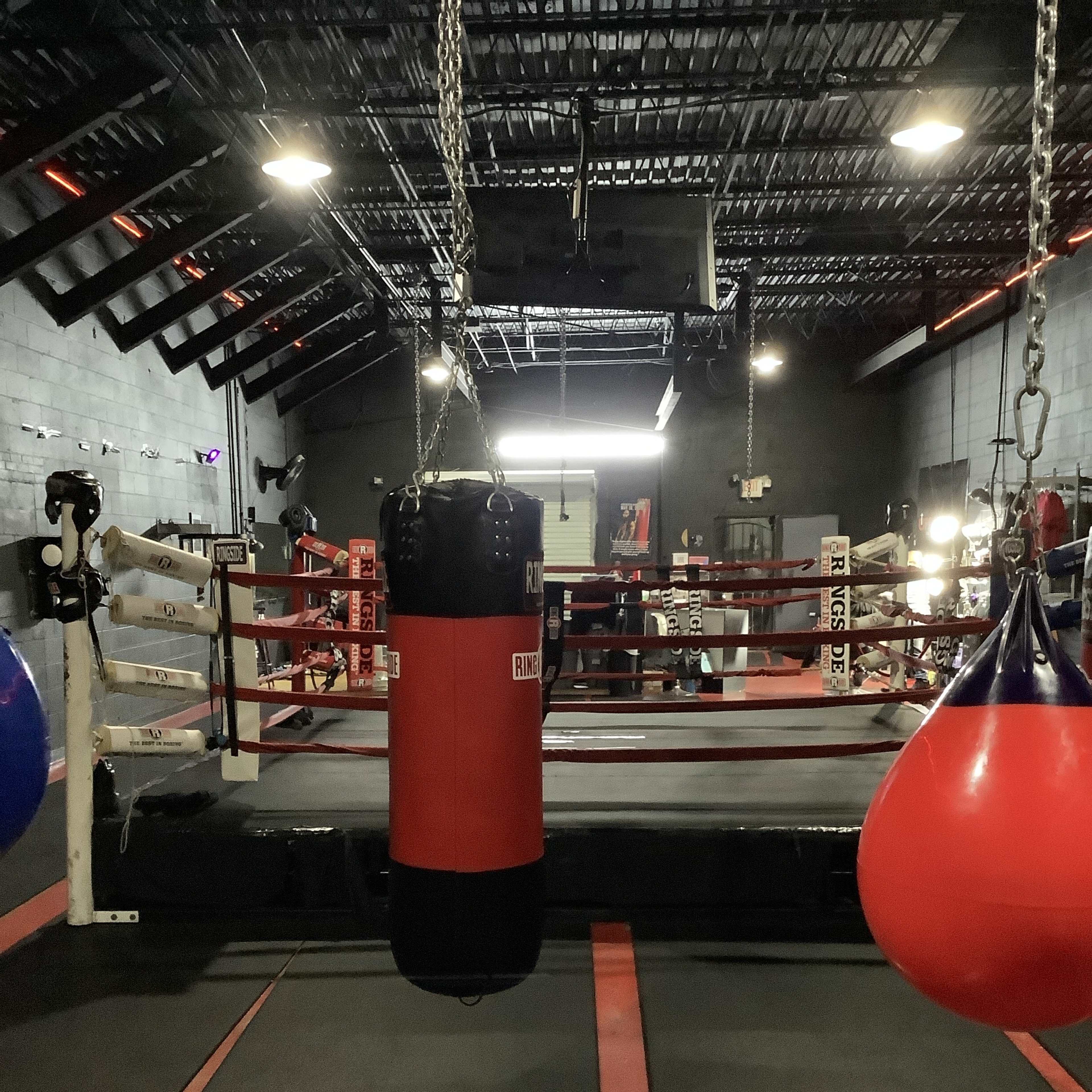 A boxing gym with a red punching bag suspended from the ceiling, surrounded by a boxing ring and exercise equipment.