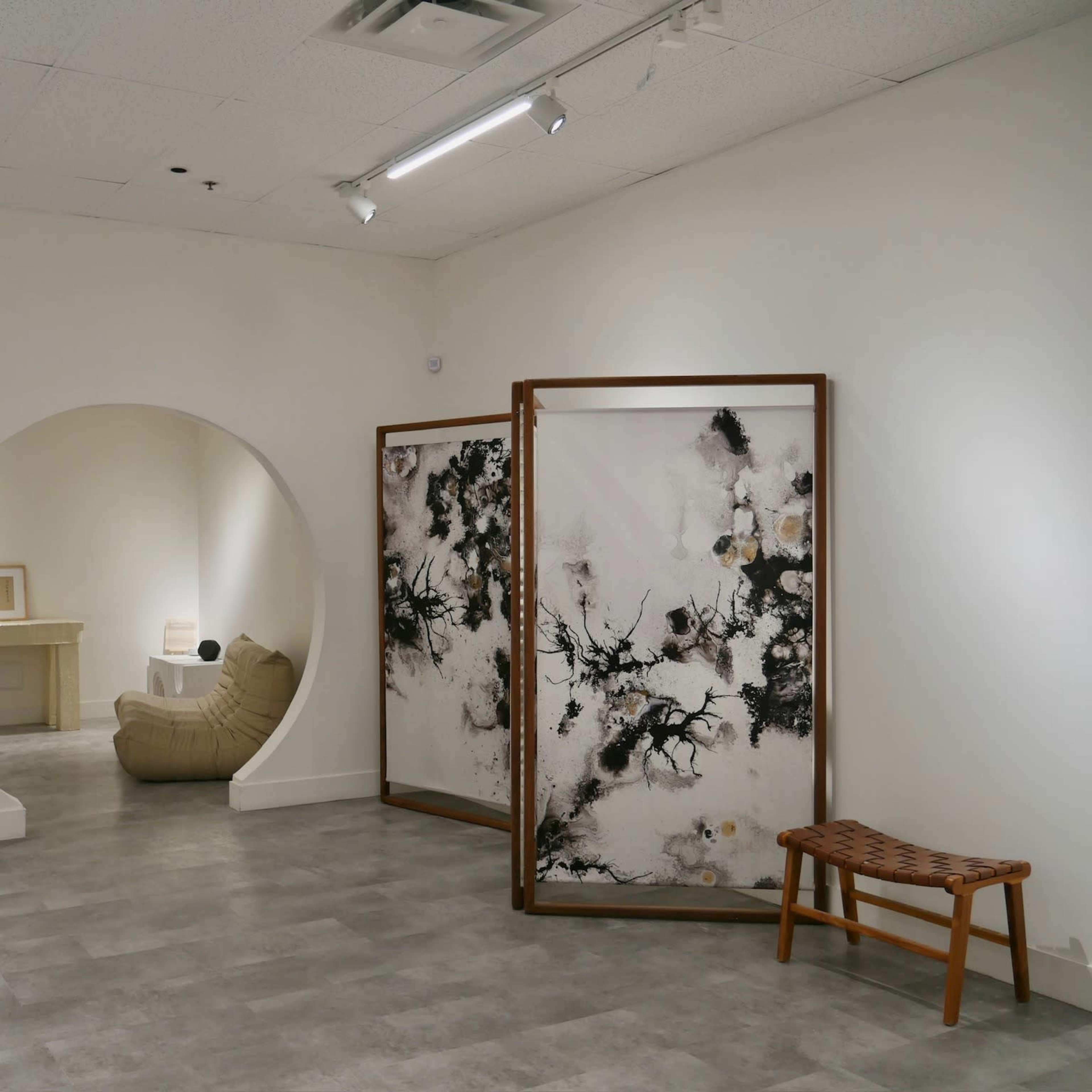 A minimalist room featuring a curved archway leading to a beige chair, a wooden folding screen with a black and white design, and two wooden benches against light-colored walls and flooring.