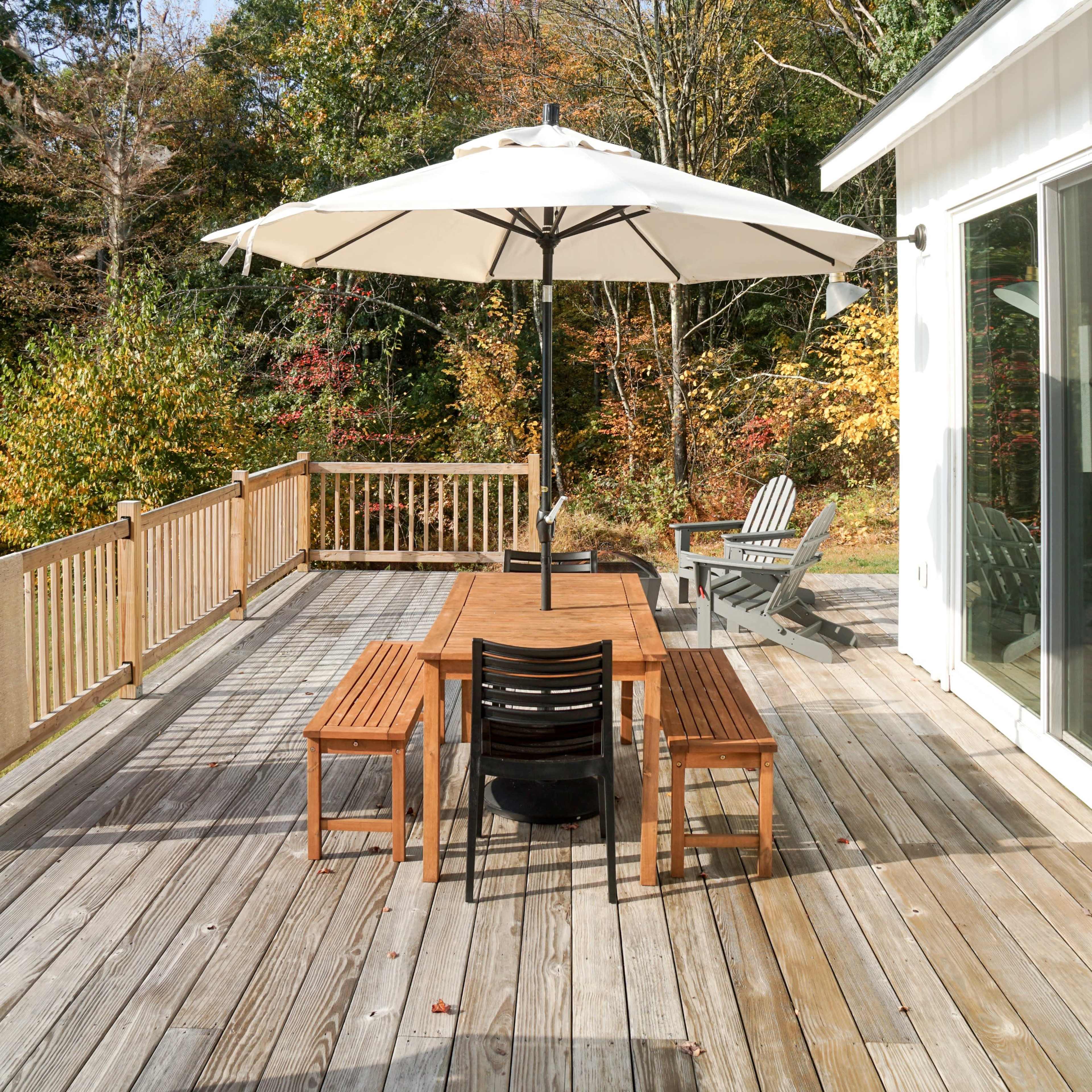 A wooden deck with a table and chairs sits beneath a large umbrella, surrounded by trees with autumn foliage.