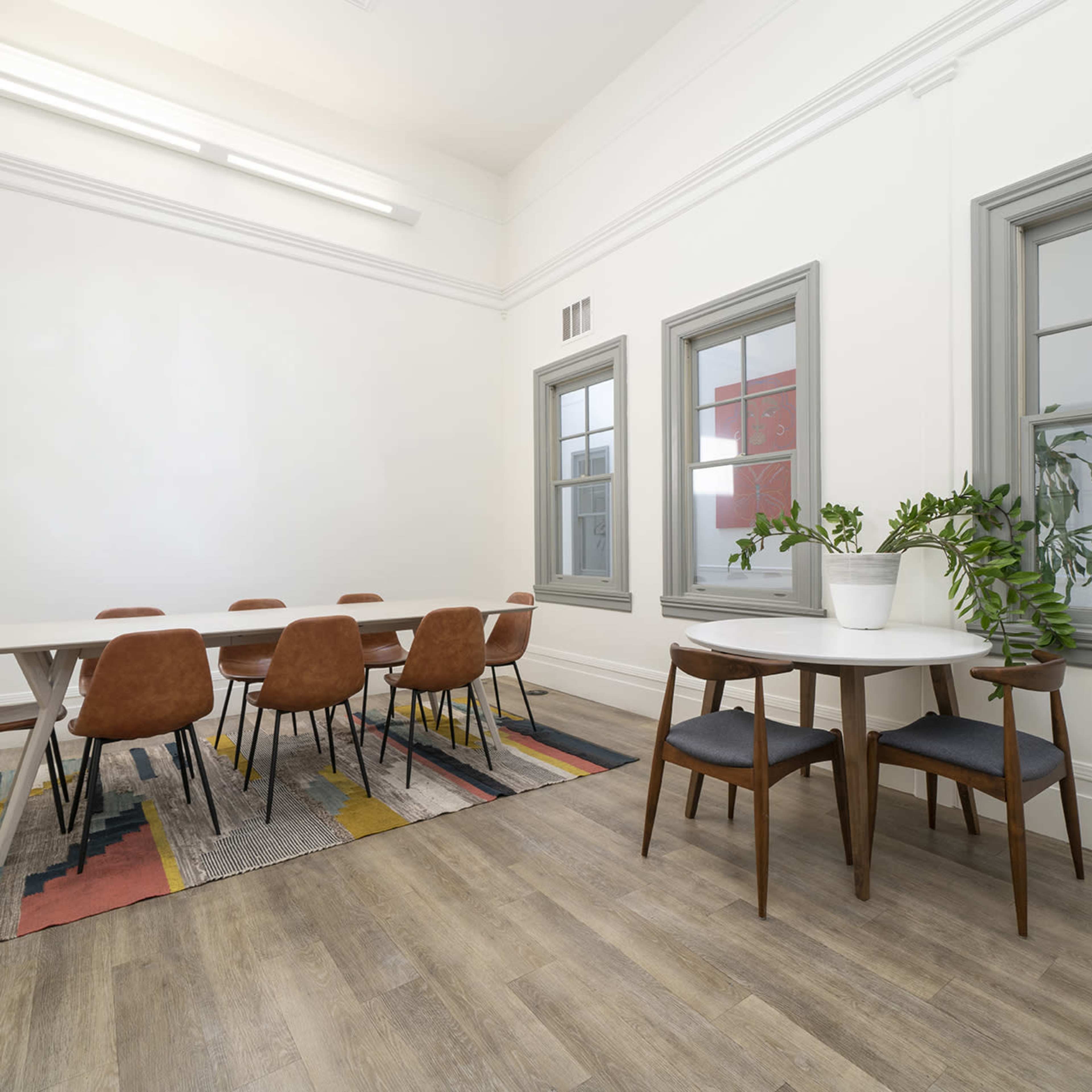 A modern meeting room features a large table surrounded by brown chairs, with a smaller round table and green plants on a patterned rug.