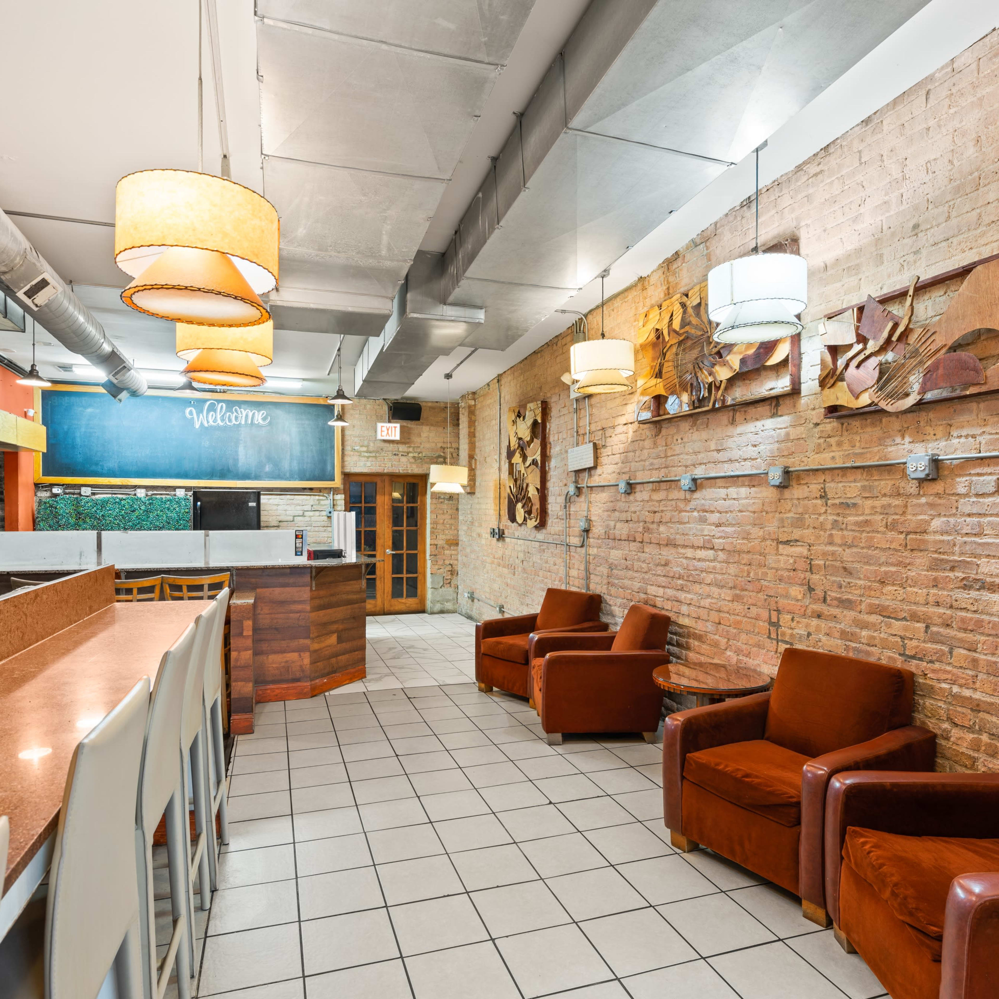 A cozy interior of a café or lounge featuring a bar counter, seating area with orange armchairs, and decorative wall art made of wood.
