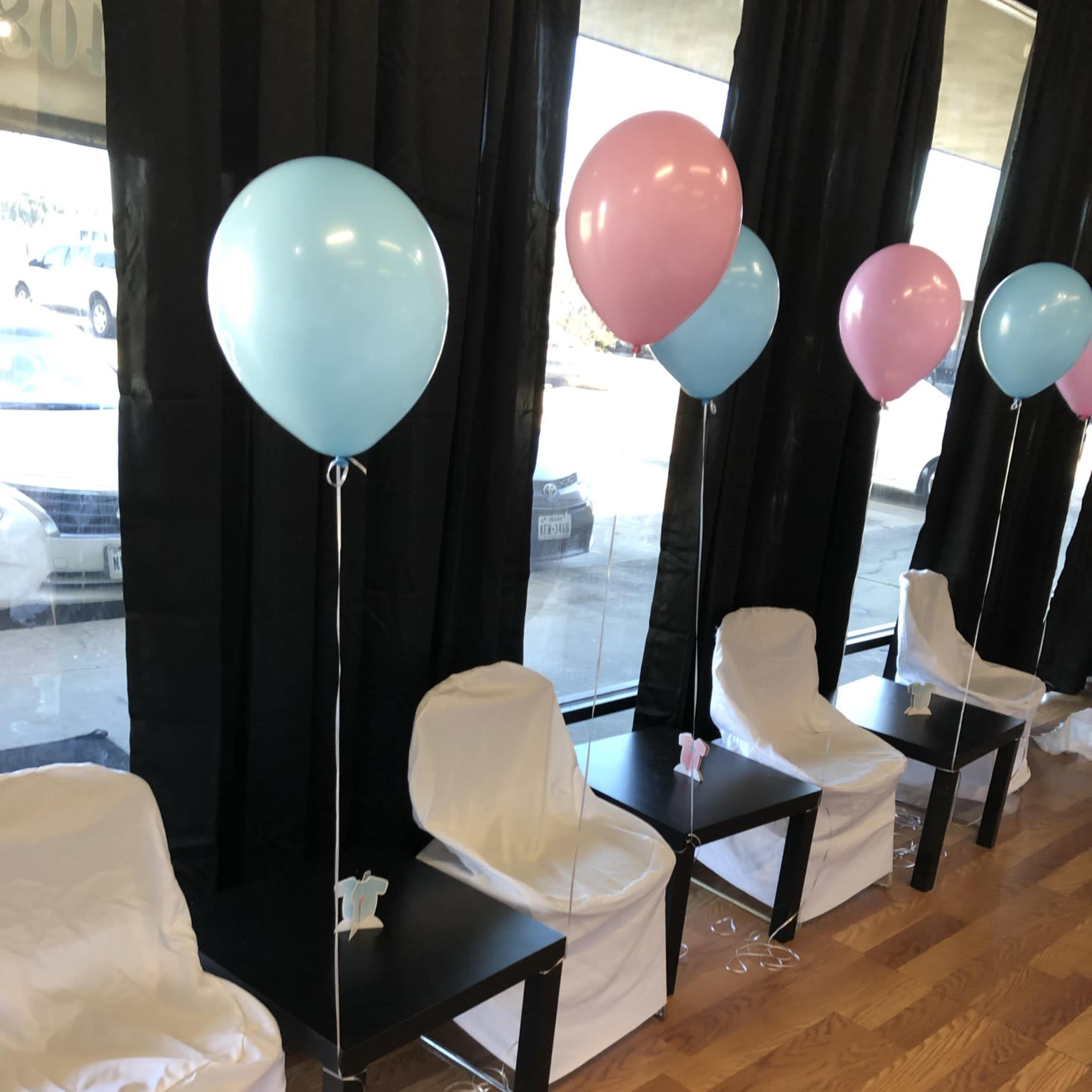 A row of four chairs covered in white cloth is set up next to a window, each adorned with pastel blue and pink balloons.