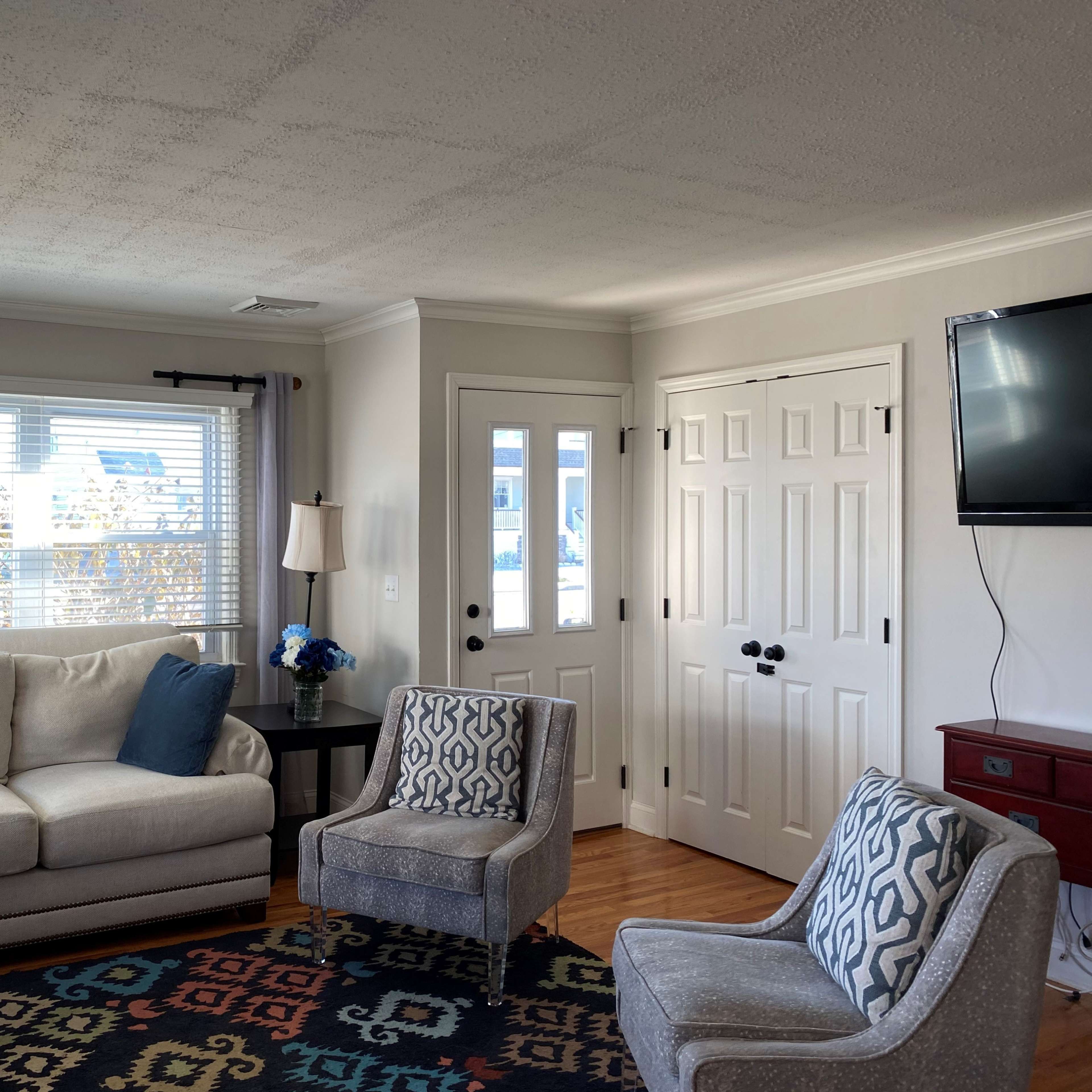 A bright living room with a light-colored sofa, two patterned chairs, a coffee table, and a television mounted on the wall, alongside a door leading outside.