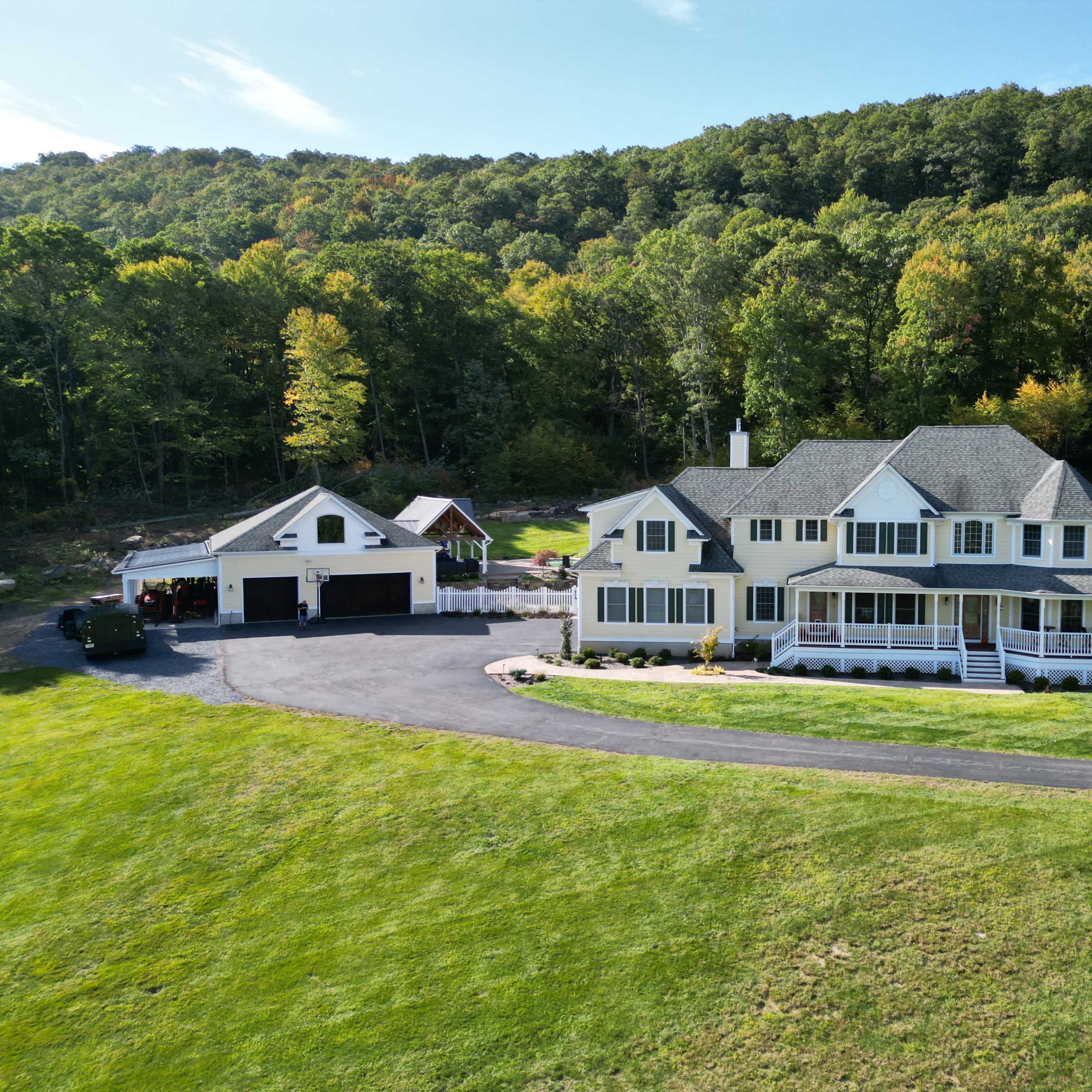 A large yellow house with a wraparound porch sits beside a garage on a landscaped property surrounded by trees.