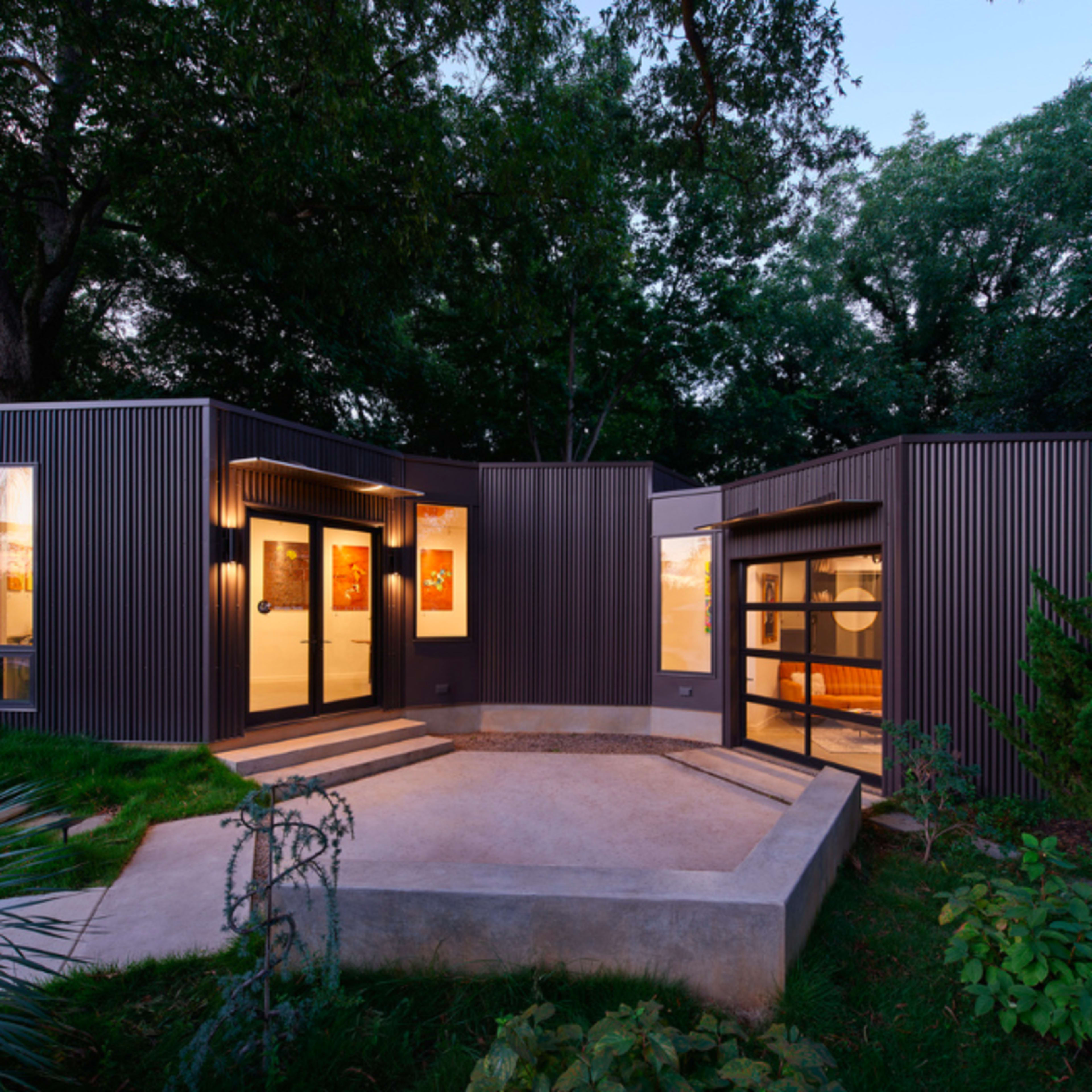 A modern, angular house with metal siding and large windows, surrounded by greenery and featuring a concrete patio.
