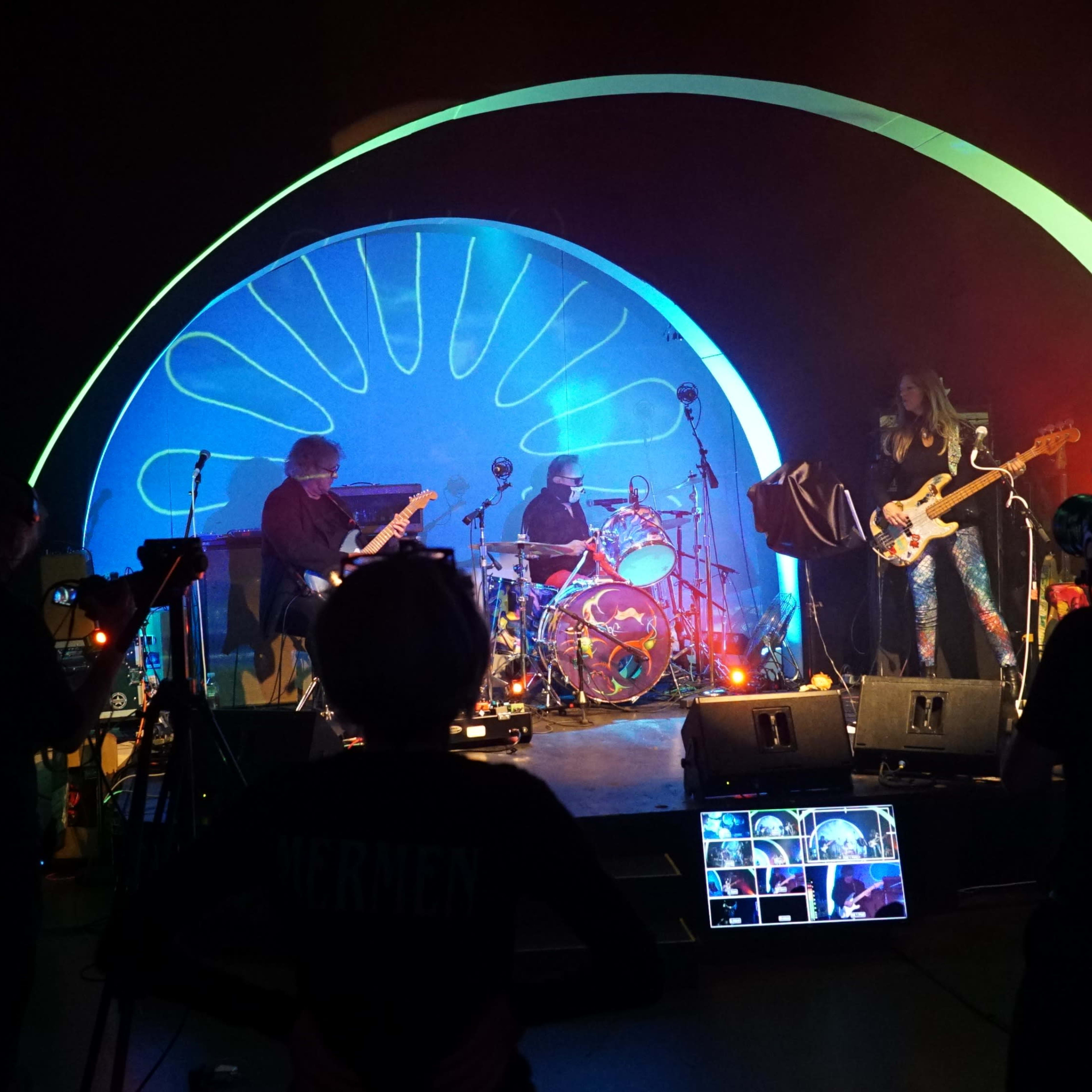 A band performs on stage with colorful lighting and a large backdrop, while cameras capture the scene.