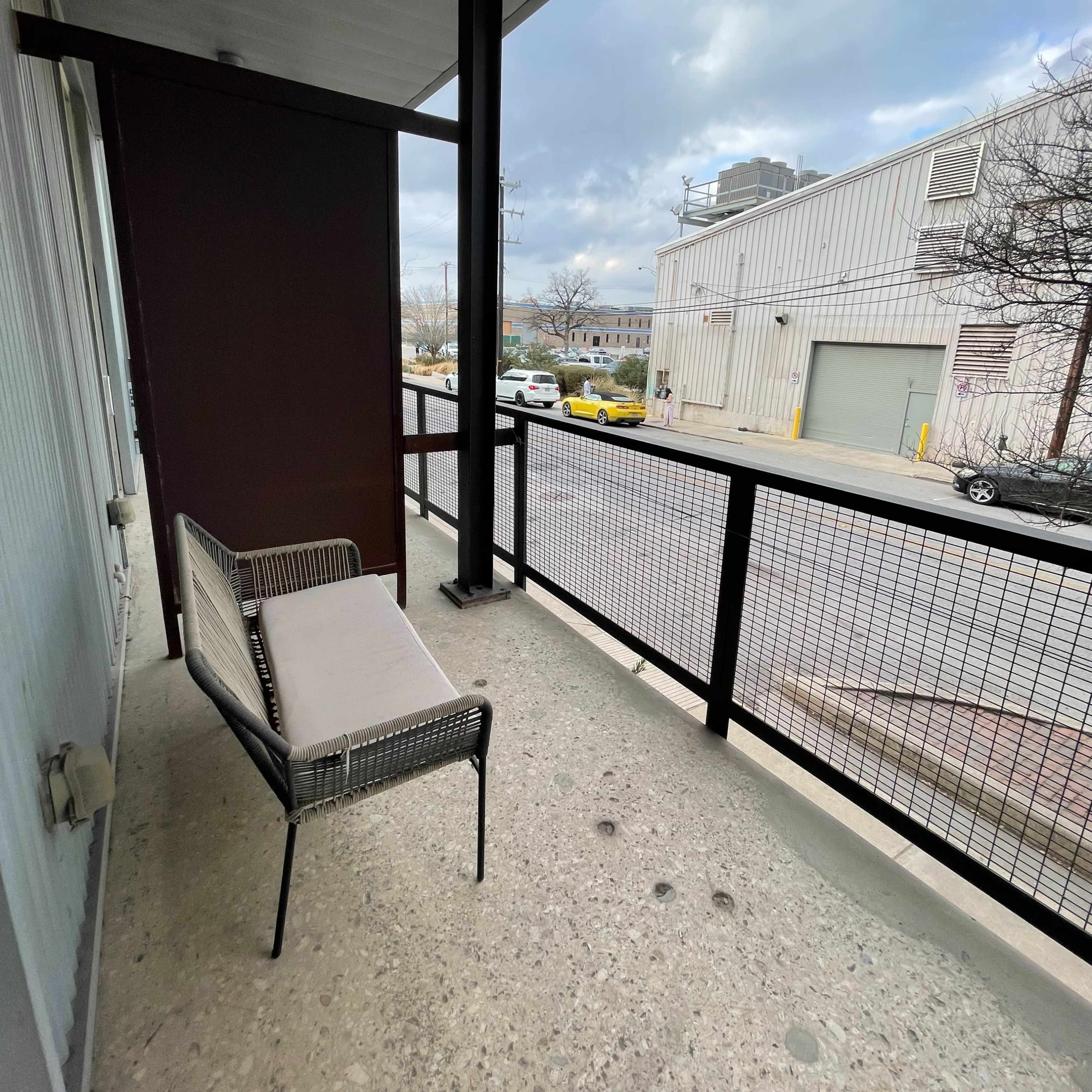 A narrow balcony with a metal-framed chair and a view of a street lined with parked cars and a building in the background.