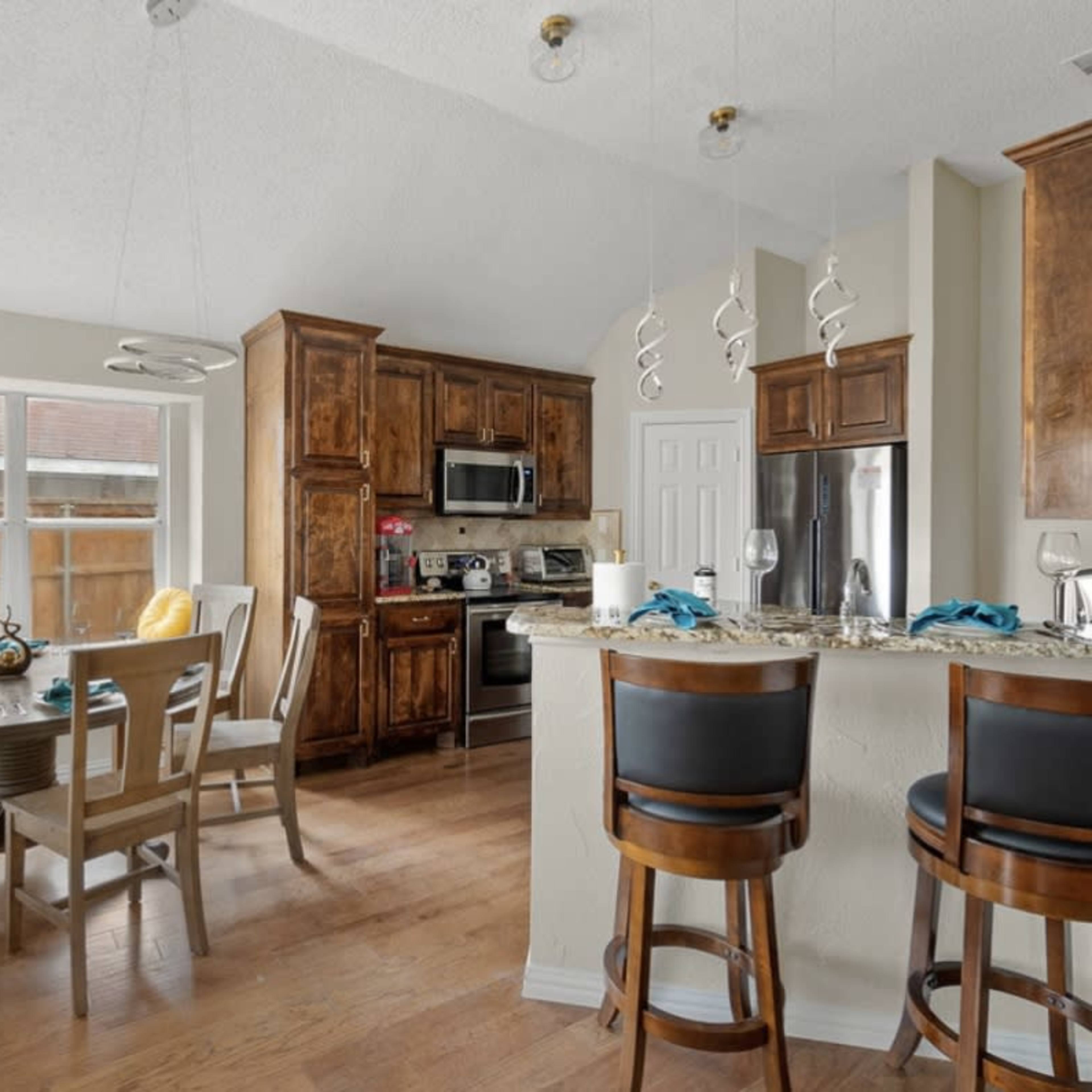 A kitchen features wooden cabinets, stainless steel appliances, and a dining area with a round table and bar stools.